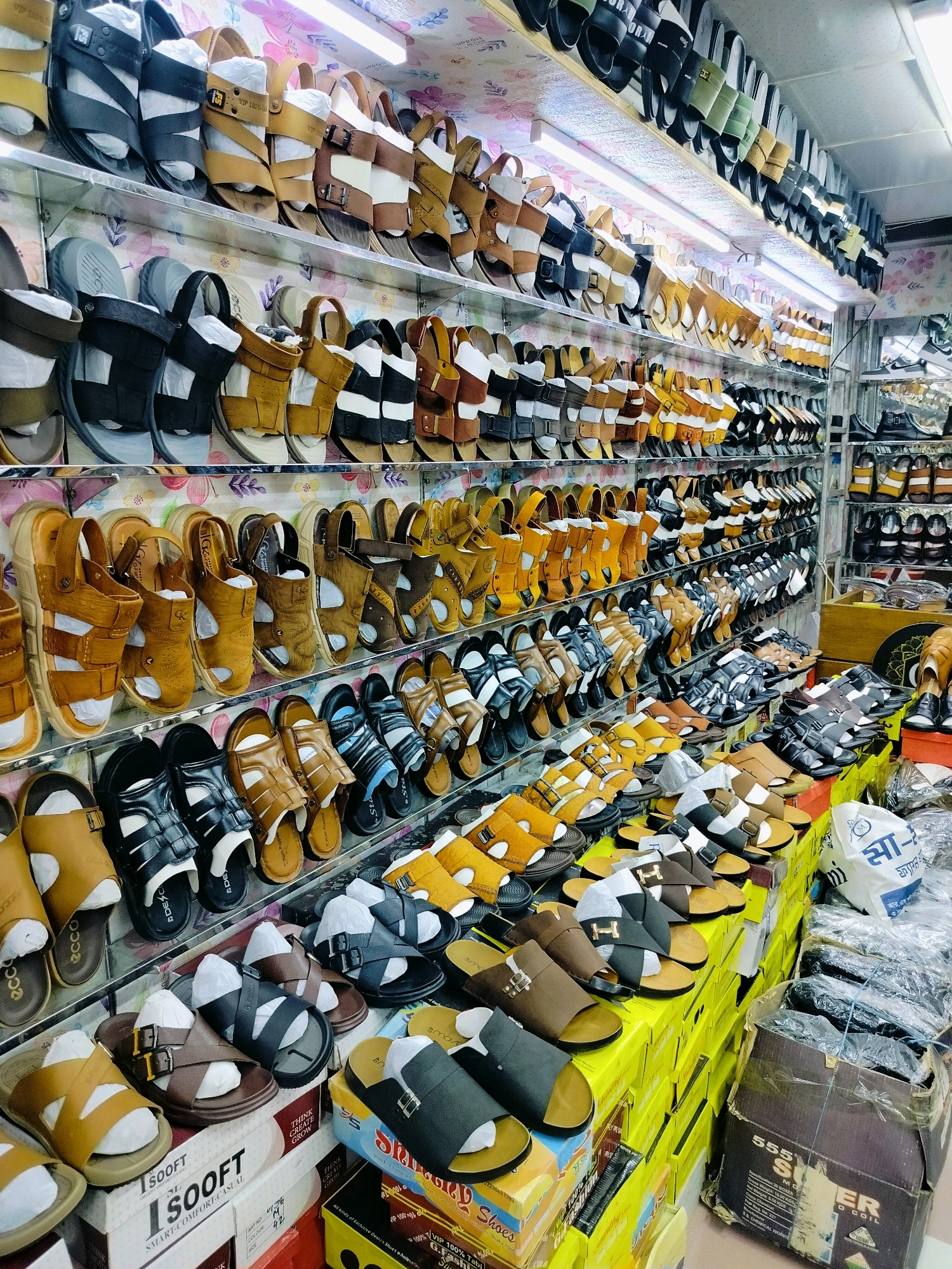 The image shows a well-stocked shoe store in Dhaka, Bangladesh, showcasing a wide variety of sandals and slippers displayed on metal shelves. The store appears to be organized with shelves meticulously filled with footwear in various colors, styles, and materials, primarily leather and synthetic. Many of the shoes are for men, featuring designs like sliders, flip-flops, and sandals with straps. Some shoes are packaged in white paper or plastic, indicating they are new. The store's interior is brightly lit by fluorescent lights, and the shelves are adorned with colorful patterned wallpaper. Boxes of shoes are stacked on the floor in front of the shelves, some of which are also visible. The overall impression is one of a busy retail environment, typical of a commercial area in Dhaka, with a focus on affordable and fashionable footwear.