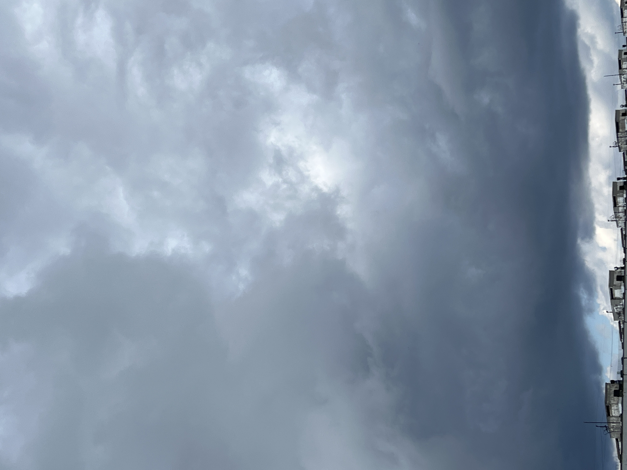The image captures a cloudy sky over the city of Tolyatti, Russia. The sky is filled with dark, heavy clouds, suggesting overcast weather. A few buildings with flat roofs and visible rooftop structures, possibly vents or small structures, are visible along the bottom edge of the frame. These buildings appear to be low-rise structures, possibly industrial or residential blocks. No people are visible in the image, and no specific actions or interactions are depicted. The overall scene is urban and somewhat subdued due to the cloudy conditions.