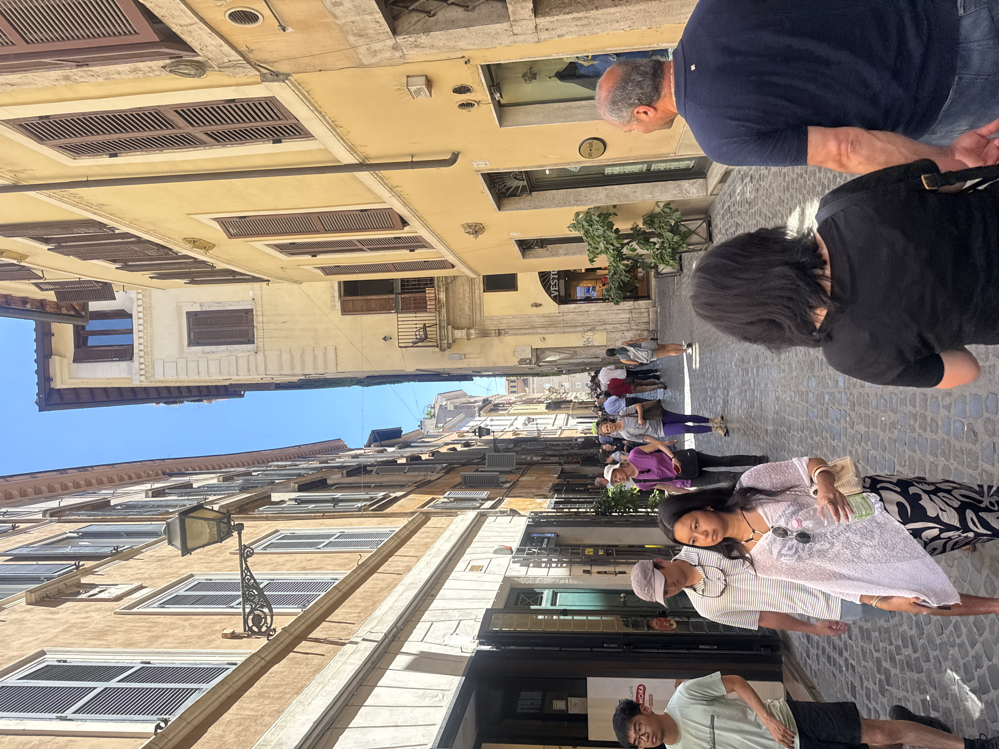 The image depicts a street scene in Rome, Italy, characterized by narrow cobblestone streets lined with buildings featuring traditional Italian architecture. The buildings are multi-storied, with windows adorned with shutters. Several people are walking along the street, suggesting a bustling urban environment. The scene is bathed in sunlight, creating distinct shadows. A street lamp is affixed to the wall. People in the frame are dressed in casual attire, typical for tourists. The overall impression is that of a vibrant, historical European cityscape.