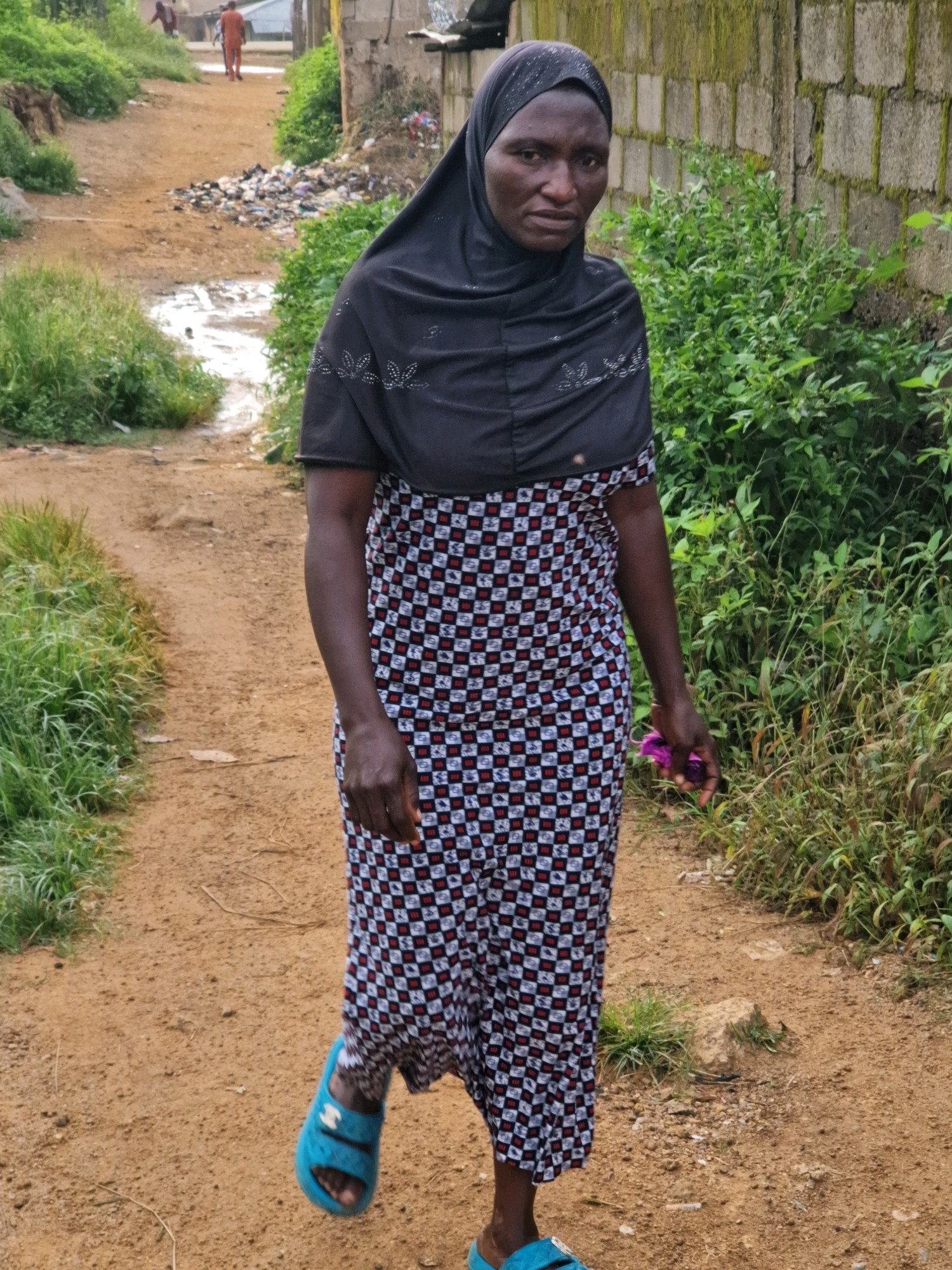 A full-body, medium shot captures a Black woman walking on a dirt path in Jos, Nigeria. The woman wears a black headscarf with white embellishments, a long, patterned dress in black, white, and red, and bright blue slides. She holds a small, purple object in her right hand, and her left hand is by her side. She is walking away from the camera, looking towards the left side of the frame. The environment is somewhat unkempt, with dense green foliage on the right side of the path and a heap of refuse in the background on the left. A brick wall is visible behind the woman on the right, and a few individuals in the distance on the path appear to be wearing orange and red clothing. The ground is a mix of dirt and small stones, with some puddles of water visible.