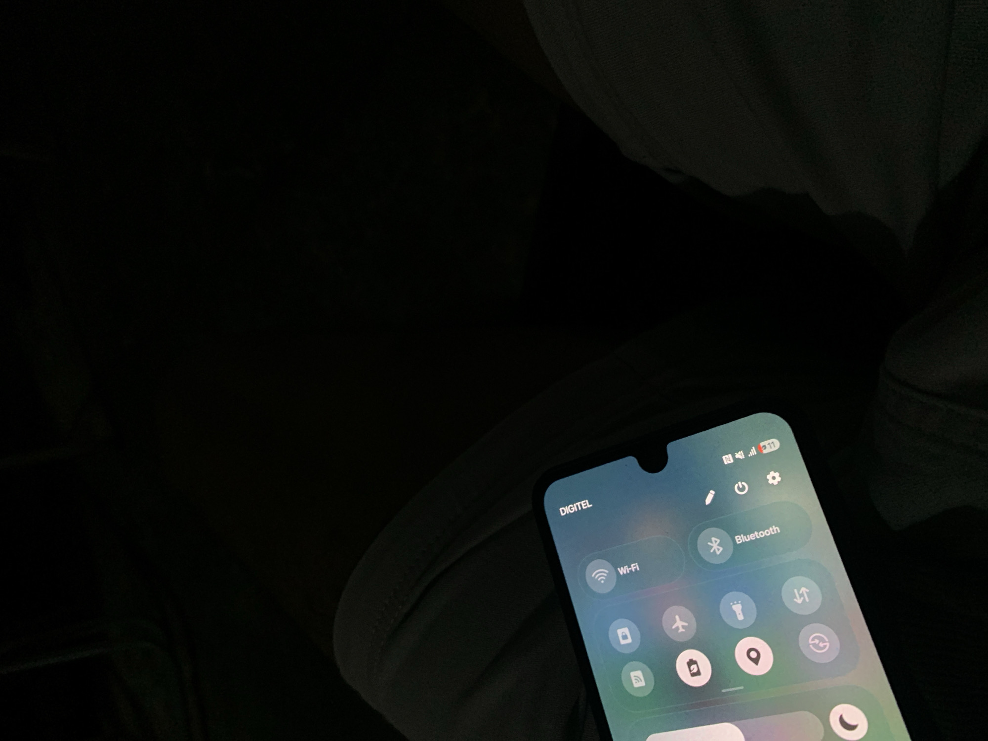The image is a close-up, low-light shot of a person's lap with a smartphone displaying a quick settings menu. The phone is angled upwards and to the right. The screen shows icons for Wi-Fi, Bluetooth, airplane mode, and flashlight, along with a brightness slider. The top right corner of the screen displays "DIGITEL", indicating a Venezuelan mobile carrier. The background is dark, with the folds of dark gray fabric visible on either side of the phone, suggesting the person is wearing pants or a similar garment. The scene is not specific to any particular location in Santa Ana, Venezuela, as it only depicts a close-up of a person and their phone.
