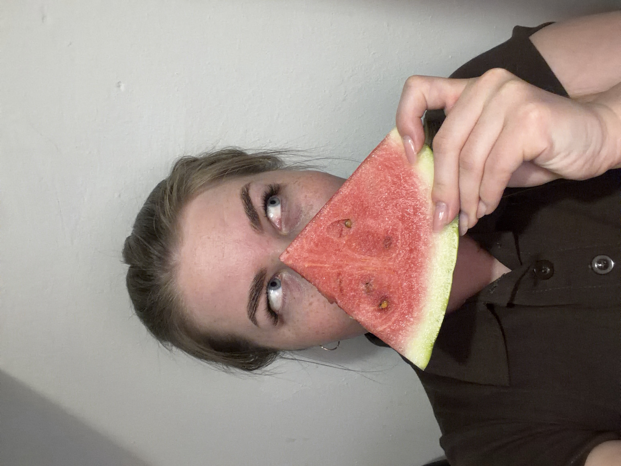 A fair-skinned person with blue eyes holds a slice of watermelon in front of their face. The watermelon obscures the center of the face. The person is wearing a dark brown collared shirt. The background is a plain, light gray wall. There is no indication of the city of Chaikovsky, Russia.