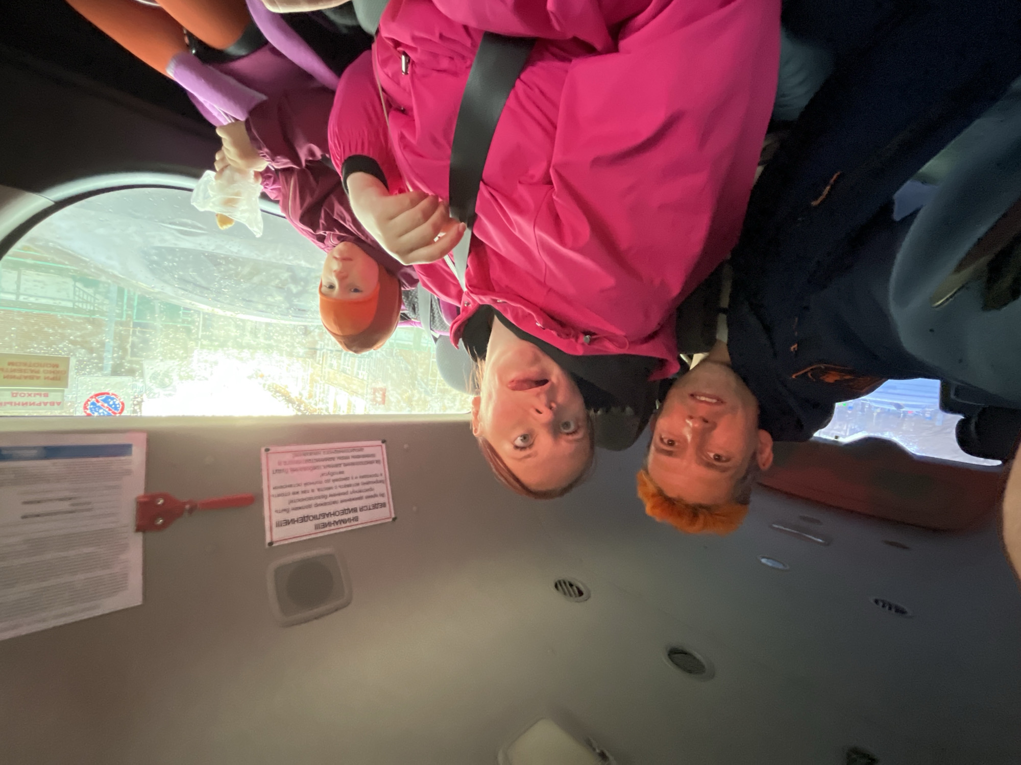 The image is presented upside down, depicting three individuals inside what appears to be a public transport vehicle, such as a bus or shuttle, likely in Izhevsk, Russia.
In the foreground, from left to right (after rotating the image), are two women and a man. The first woman on the left, partially visible, is wearing an orange hat and a maroon jacket. She holds a plastic bag containing an item. The second woman in the center is wearing a bright pink jacket with a black seatbelt across her chest and a black top underneath. She is looking directly at the camera with a playful, surprised expression, mouth open with her tongue slightly out, and her eyes wide and looking slightly upward. She has reddish-brown hair. To her right is a man with short, bright orange-red hair, wearing a dark blue top or jacket. He is also looking at the camera with a slight smile.
The bright outdoor light streams through a window on the left, which appears wet, suggesting rain or snow. Reflections are visible on the window surface. The background shows the vehicle's interior, including parts of seats (one appears orange/red, another dark blue), ceiling vents, and lighting fixtures.
Several official-looking notices and a red emergency hammer are affixed to the interior wall of the vehicle. Visible text on the right sign (translated from Russian) reads: "ATTENTION!!! VIDEO SURVEILLANCE!!! VIDEO IS BEING CONDUCTED..." followed by instructions that a "passenger must be belted with a seatbelt!" and mentions of "also stand" and "stop of the bus." Another partially visible sign on the left mentions "EMERGENCY," "EXIT," and "WITH A HAMMER," likely indicating emergency exit procedures involving breaking a window with the hammer.
The subjects' warm clothing (jackets, hats) suggests cooler weather conditions. The overall atmosphere conveyed by their expressions is lighthearted and candid.