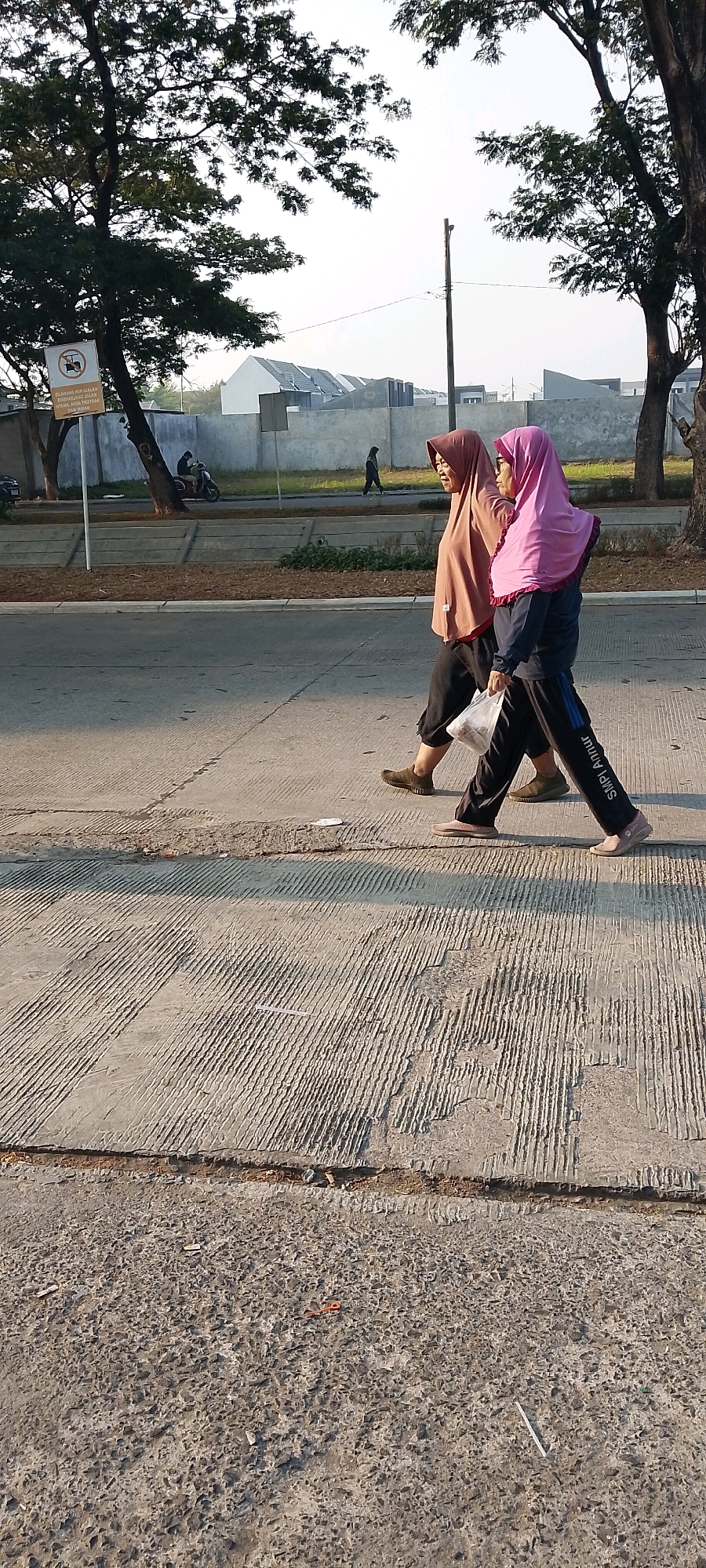 Two women are walking on a paved path in what appears to be a suburban area of Satriamekar, Indonesia. The woman on the left is wearing a brown hijab and a loose brown shirt with dark pants. The woman on the right is wearing a pink hijab, a dark blue long-sleeved shirt, and dark pants with white writing on the left leg. She is carrying a clear plastic bag filled with white items. They are walking side-by-side, and the woman on the right is looking forward while the woman on the left is looking slightly to her right. The path is textured concrete, with some areas appearing to have been repaired. In the background, there is a road with a few vehicles, a concrete barrier, a wall with a sign, and the construction of residential buildings with white roofs. Trees with green foliage are present on the left and right sides of the image. The sky is bright and clear, suggesting it is daytime.