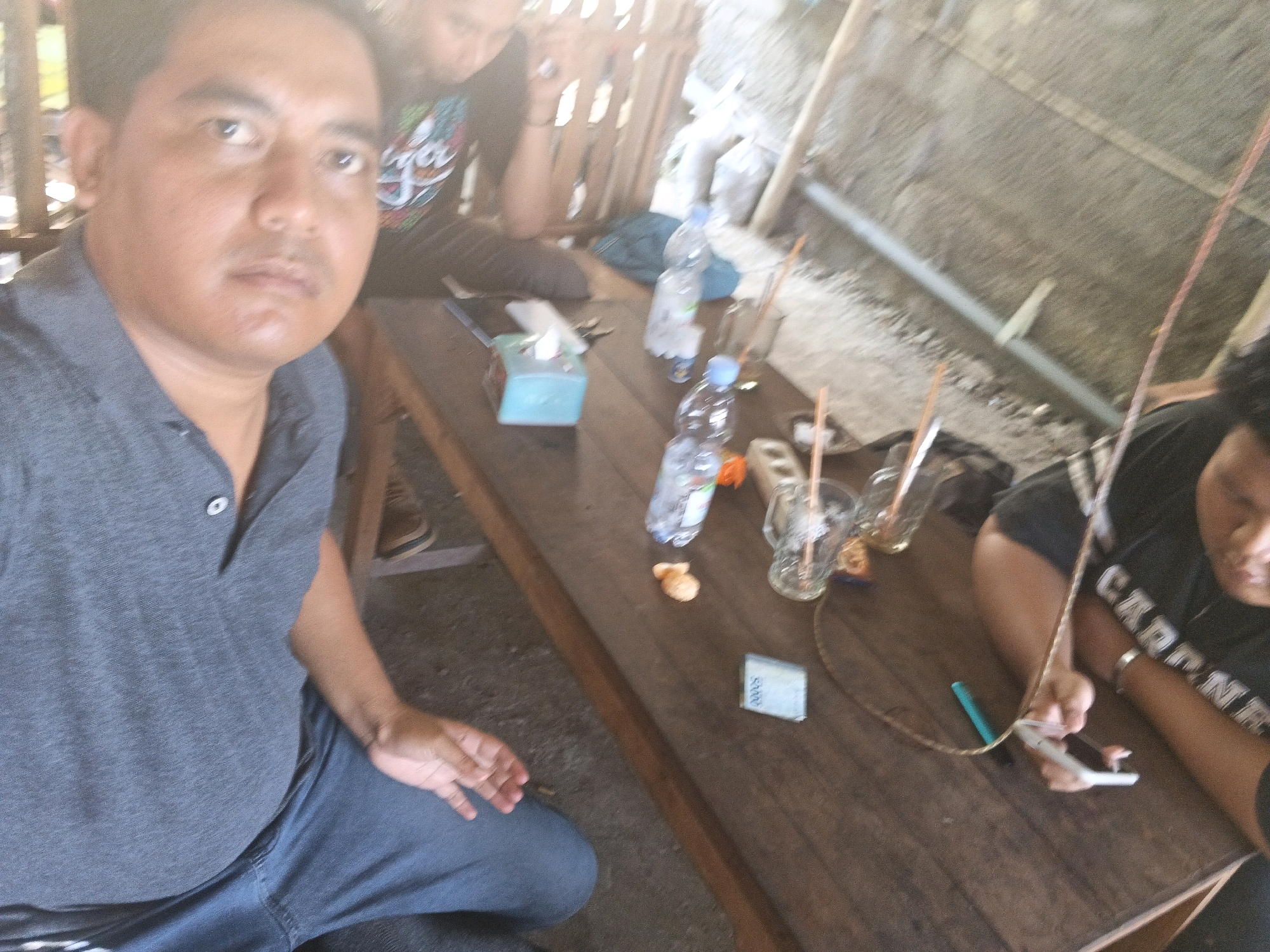 The image depicts a gathering of at least three individuals seated around a wooden table, likely in an outdoor or semi-outdoor setting. The scene appears to be casual, possibly a roadside eatery or a local gathering spot in Kecamatan Pati, Indonesia. In the foreground, a man wearing a grey polo shirt and blue jeans is prominently featured, looking directly at the camera with a neutral expression. His left arm rests on his leg. To his right, on the table, are several items including a tissue box, a bottle of water, and a small orange piece of food. Further to the right, another individual, partially obscured, is seen from the chest up, wearing a dark t-shirt with white lettering that reads "CARNIVORES". This person is holding a mobile phone. Behind them, another person is visible, also wearing a dark shirt with graphics. The table is a rustic wooden surface, cluttered with bottles, glasses with straws, and a piece of what appears to be currency, likely Indonesian Rupiah. The background is blurred, suggesting a shallow depth of field, but glimpses of bamboo-like structures and roofing materials indicate a simple, possibly rustic building. The overall lighting is soft, with some areas appearing overexposed, typical of natural light conditions. The environment suggests a relaxed and informal social interaction.