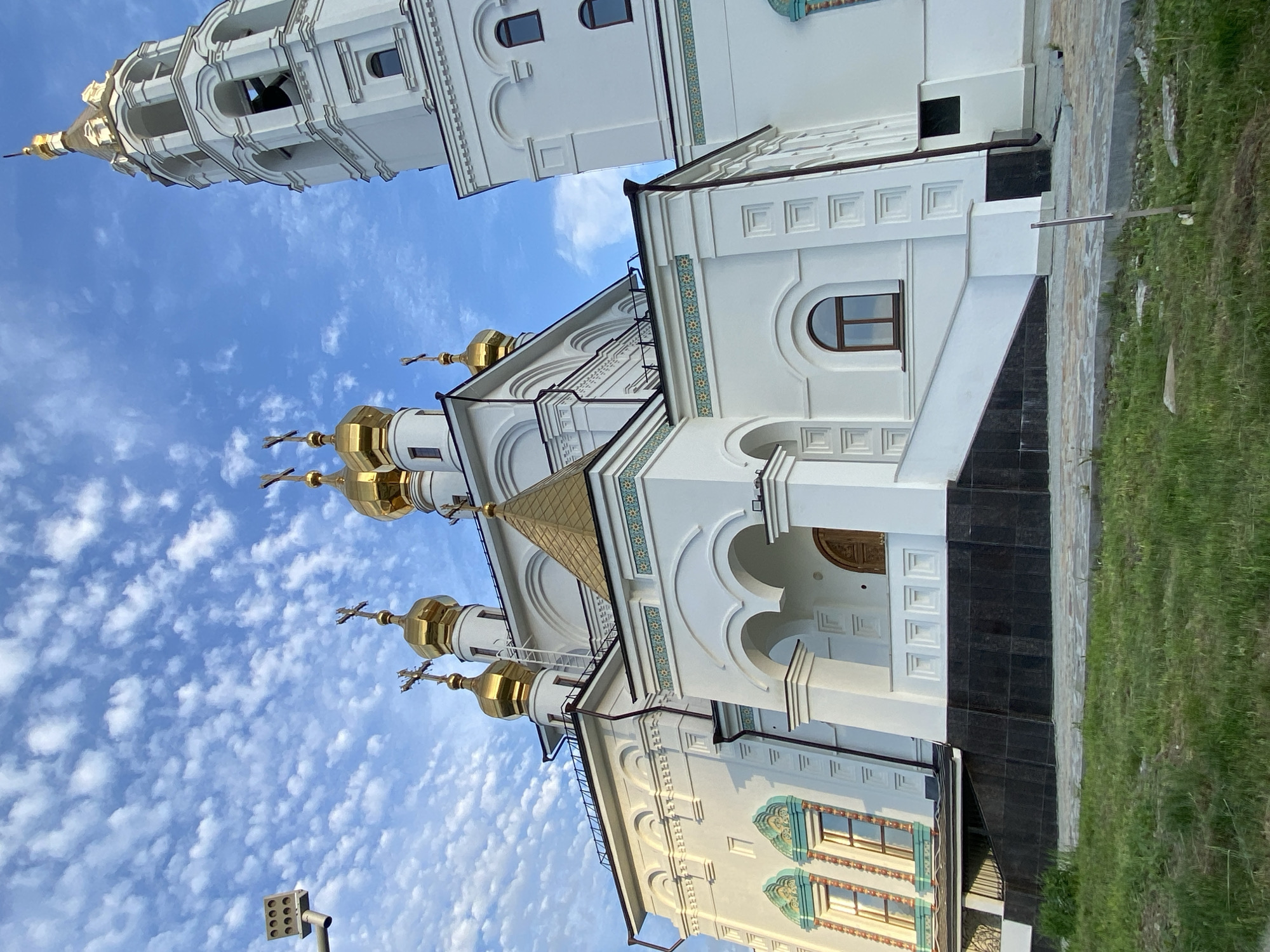The image depicts a white Orthodox church building with golden domes and crosses against a blue sky with scattered clouds. To the left is a tall, white bell tower. The church's architecture includes arched windows and decorative tilework. The setting appears to be outdoors, with grass visible at the bottom of the frame. Based on the architecture, it is likely the Church on Blood in Yekaterinburg, Russia.