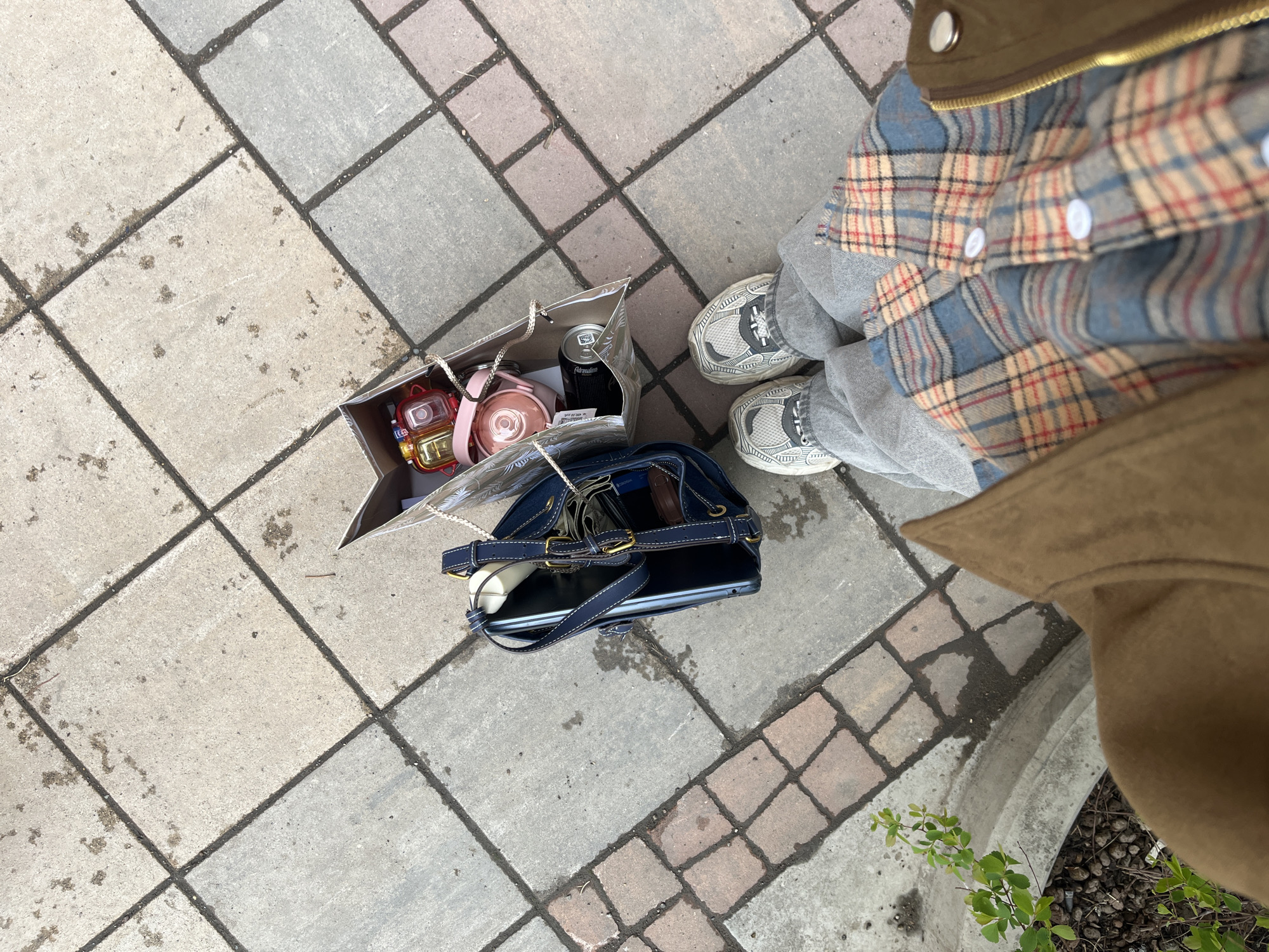 The image is a high-angle shot of a person standing on a paved surface, likely a sidewalk or pedestrian area. The pavement consists of large, irregularly shaped concrete or stone tiles in varying shades of gray and pink, separated by dark grout lines. The person is wearing gray sneakers, gray sweatpants, and a blue and tan plaid shirt under a tan jacket. A brown paper shopping bag sits on the pavement to the left of the person. The bag contains various items, including a pink thermos, a black can (likely a beverage), and other smaller, unidentified objects. A dark blue purse with a black laptop inside is on the pavement in front of the shopping bag. In the bottom right corner, a small patch of green plants is visible, possibly growing in a nearby planter. The location cannot be determined from the image provided.