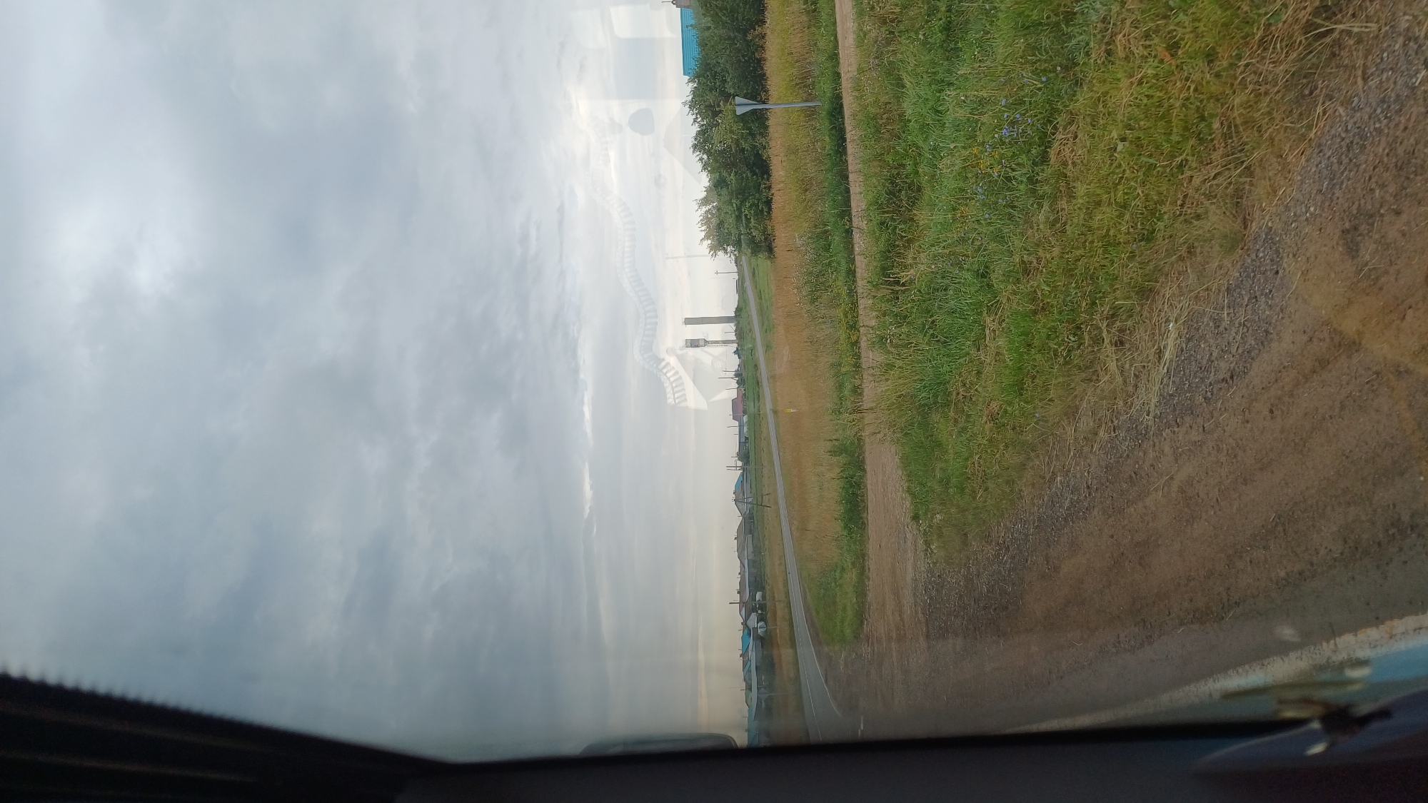 The image depicts a cloudy, overcast day, likely in the village of Kiyasovo, Russia, as suggested by the presence of traditional Russian architecture and a rural landscape. The scene is viewed from inside a vehicle, indicated by the reflection on the window.
In the mid-ground, a paved road curves to the left, bordered by a grassy verge. Beyond the road, a cluster of low-rise houses with multi-colored roofs, typical of rural Russian settlements, stretches across the landscape. Some of these houses appear to be of wood construction. In the distance, two tall, cylindrical structures, possibly water towers or industrial chimneys, rise against the sky. The surrounding vegetation consists of fields of dry grass and scattered bushes. The foreground is dominated by the dirt and gravel shoulder of the road, with more green grass and wildflowers visible to the right. The sky is filled with a uniform layer of gray clouds.