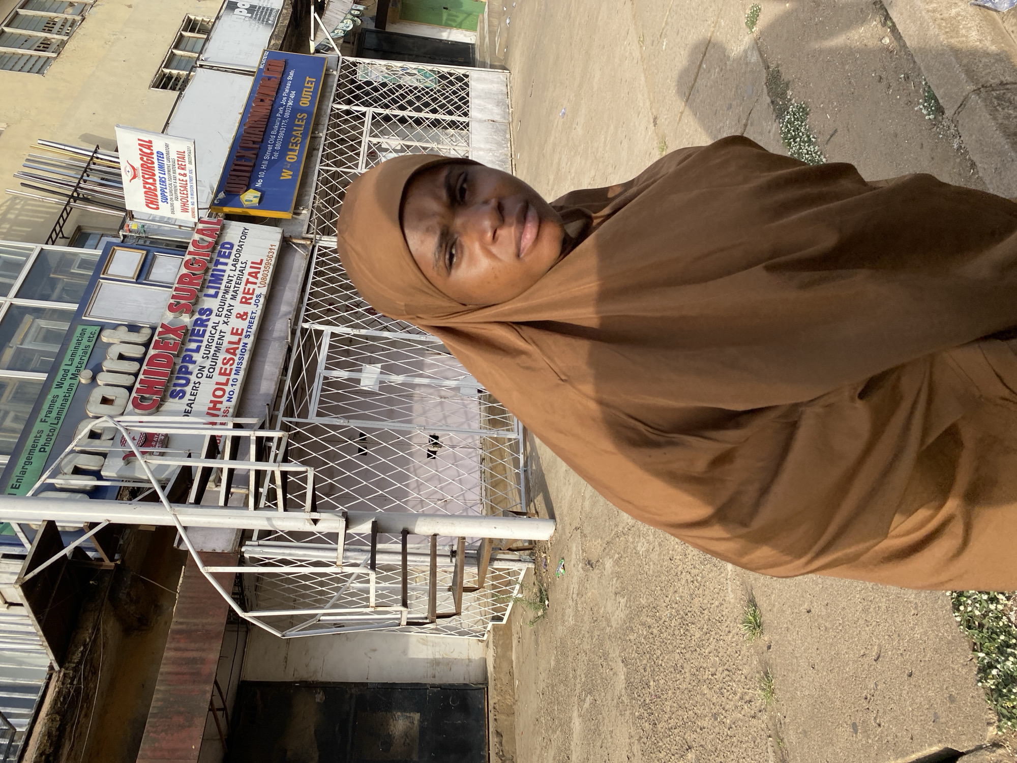 A medium shot of a woman wearing a brown hijab, standing on a concrete sidewalk. Behind her is a building with multiple signs, one of which reads "CHIDEX SURGICAL SUPPLIERS LIMITED, DEALERS ON SURGICAL EQUIPMENT, LABORATORY EQUIPMENT, X-RAY MATERIALS, WHOLESALE & RETAIL, ADDRESS: NO. 10 MISSION STREET JOS 08035956311." Another sign mentions "Hill Street Old Bukuru Park, Jos Plateau State." A metal spiral staircase is in front of the building. The lighting suggests it's daytime. Small white flowers grow between the sidewalk and the building.