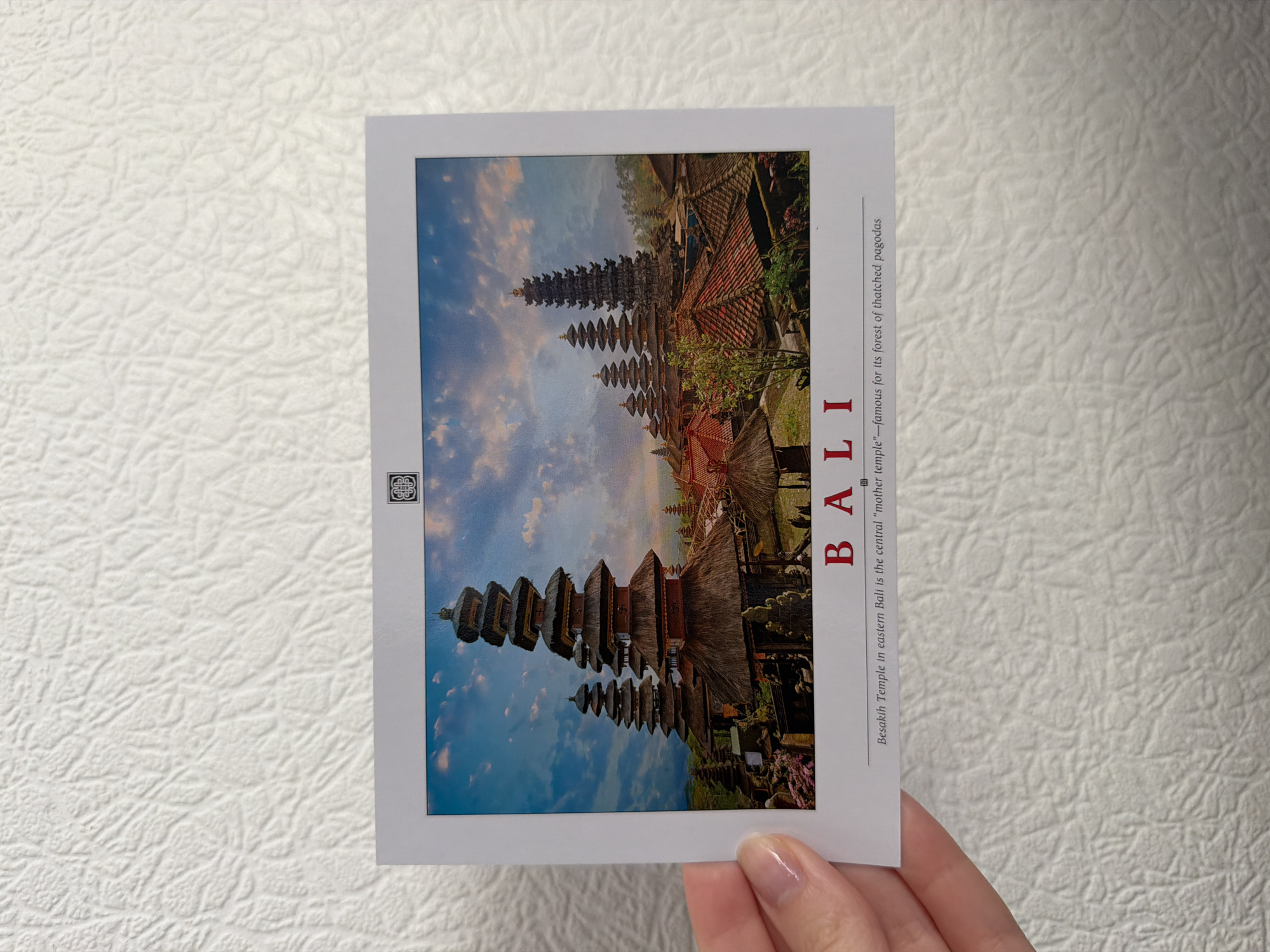 A postcard displaying a panoramic view of several multi-tiered pagodas with thatched roofs is held by a person's hand in the lower left corner. The pagodas are situated on a hillside under a dramatic sky filled with clouds. The foreground of the postcard features the word "BALI" in large, red capital letters, followed by smaller text that reads "Besakih Temple in eastern Bali is the central 'mother temple'—famous for its forest of thatched pagodas." The background of the postcard is a textured white surface, possibly a wall or a refrigerator. The postcard itself is white with a thin gray border.