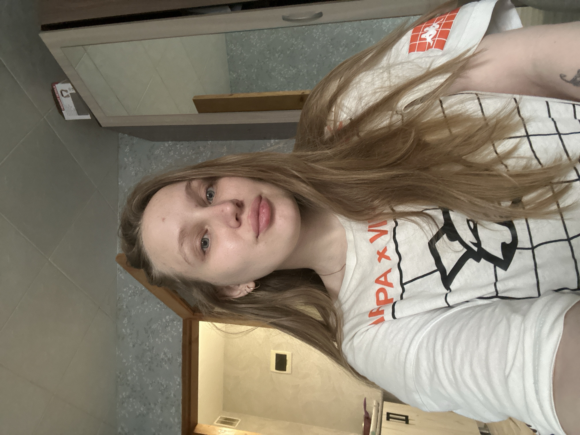 A young woman with light brown hair is facing the camera, wearing a white long-sleeved shirt with a black grid pattern and orange lettering. She appears to be in a bathroom, evidenced by the tiled walls, vanity with a mirror, and doorway leading to another room. The location is Chaikovsky, Russia, based on the metadata associated with the image.