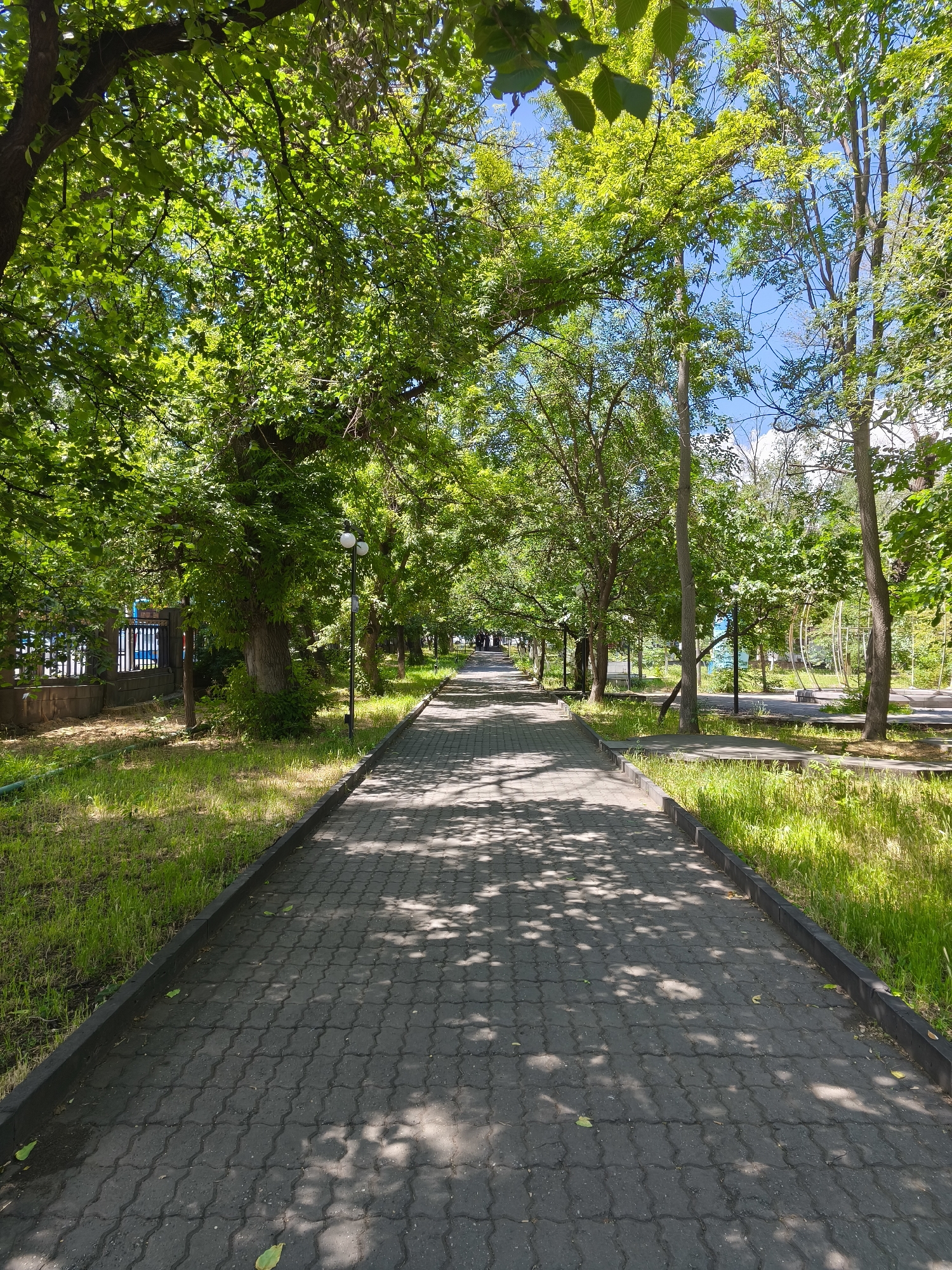 A sun-dappled pedestrian walkway, paved with interlocking gray concrete blocks, stretches into the distance. Lush green grass flanks both sides of the pathway, with mature trees forming a canopy overhead, casting intricate shadows onto the ground. The trees are deciduous, with vibrant green foliage characteristic of spring or summer. On the left side, a dark metal fence with vertical bars separates the walkway from a building or property. A lamppost with two white globe lights stands to the right of the path, approximately midway into the visible frame. In the background, more trees and hints of urban infrastructure, such as a playground-like structure with curved metal elements, are visible under a bright blue sky with scattered white clouds. The image appears to be taken on a sunny day in a park or along a tree-lined street in an urban setting, possibly Yerevan, Armenia, given the architectural elements and vegetation. There are no people or animals present in the scene.
