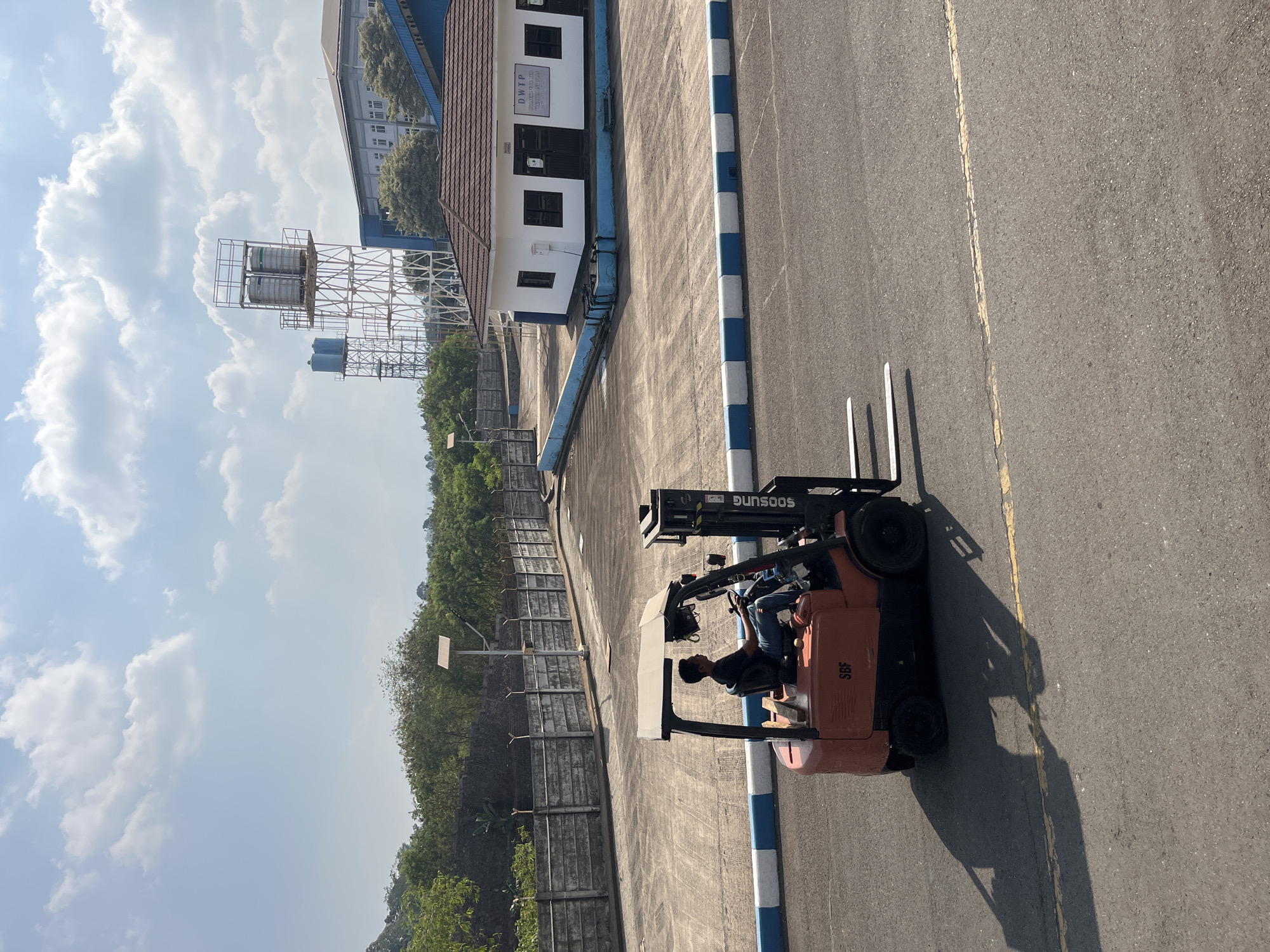 In Pelang, Indonesia, under a bright, partly cloudy sky, a man operates an orange forklift on an asphalt road. The forklift, with the brand name "SOOSUNG" visible, has its forks extended and is angled to the right of the frame. The man, wearing a dark t-shirt and blue jeans, is seated and looking forward. The forklift's shadow stretches across the road. In the background, a white building with a brown tiled roof and blue trim is visible, identified by a sign as "DWTP" (Disediakan Untuk Anda Pasti). Adjacent to the building is a large, fenced area. Further back, a tall structure with multiple water tanks stands against a backdrop of lush green trees and a distant hillside. A concrete wall with barbed wire runs along the edge of the road.