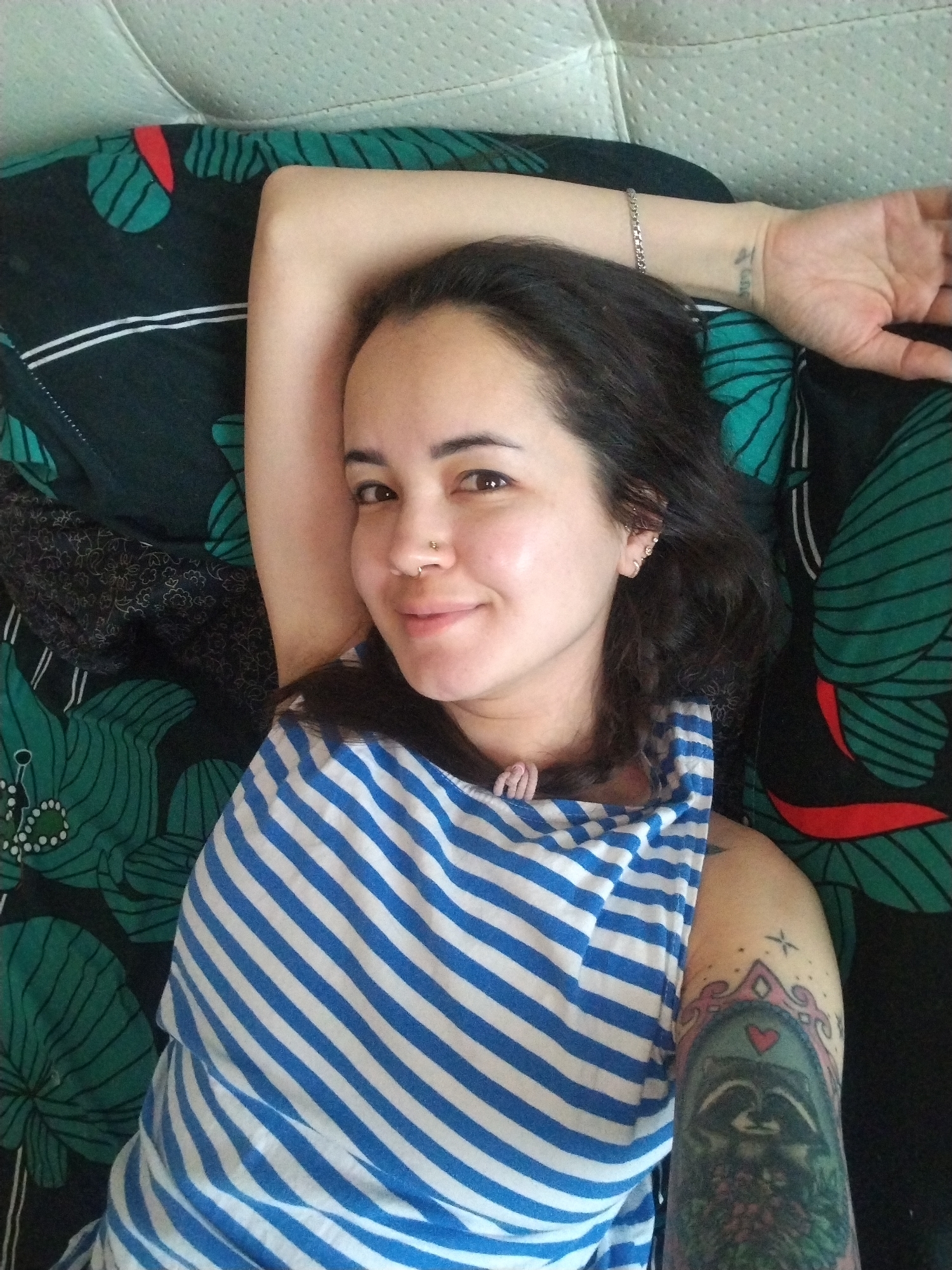 A young woman with dark hair and brown eyes lies in a relaxed pose, looking directly at the camera with a soft smile. She wears a blue and white striped tank top. Her left arm is extended above her head, showcasing a silver chain bracelet and a small, unreadable text tattoo on her inner wrist. Her right upper arm features a prominent, colorful tattoo of two raccoons enclosed within an ornate, mirror-like frame, crowned with a small red heart and surrounded by floral elements; a subtle star-like design is visible above the frame. Facial piercings include a stud in her right nostril, and multiple piercings adorn her left ear, including helix piercings. She is resting on a dark green fabric patterned with large, stylized green leaves and some red accents. Behind her head is a light-colored, padded surface with a textured pattern, likely a headboard or cushion. The image conveys a relaxed, candid moment indoors, under soft daytime lighting, potentially in Noviy, Russia.