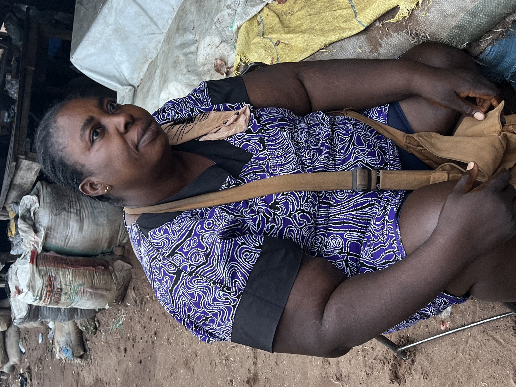 A dark-skinned woman, likely in her late 30s to 40s, is sitting outdoors in Nkpor, Nigeria. She is wearing a purple and white patterned top with black sleeves and a brown strap across her chest. She has dark hair braided and pulled back. Her hands are resting on her lap, and she appears to be holding a tan-colored bag. In the background, there are several large sacks, some of which are white and others appear to be burlap or similar material. The ground is dirt and sand. The overall impression is that of a market scene.