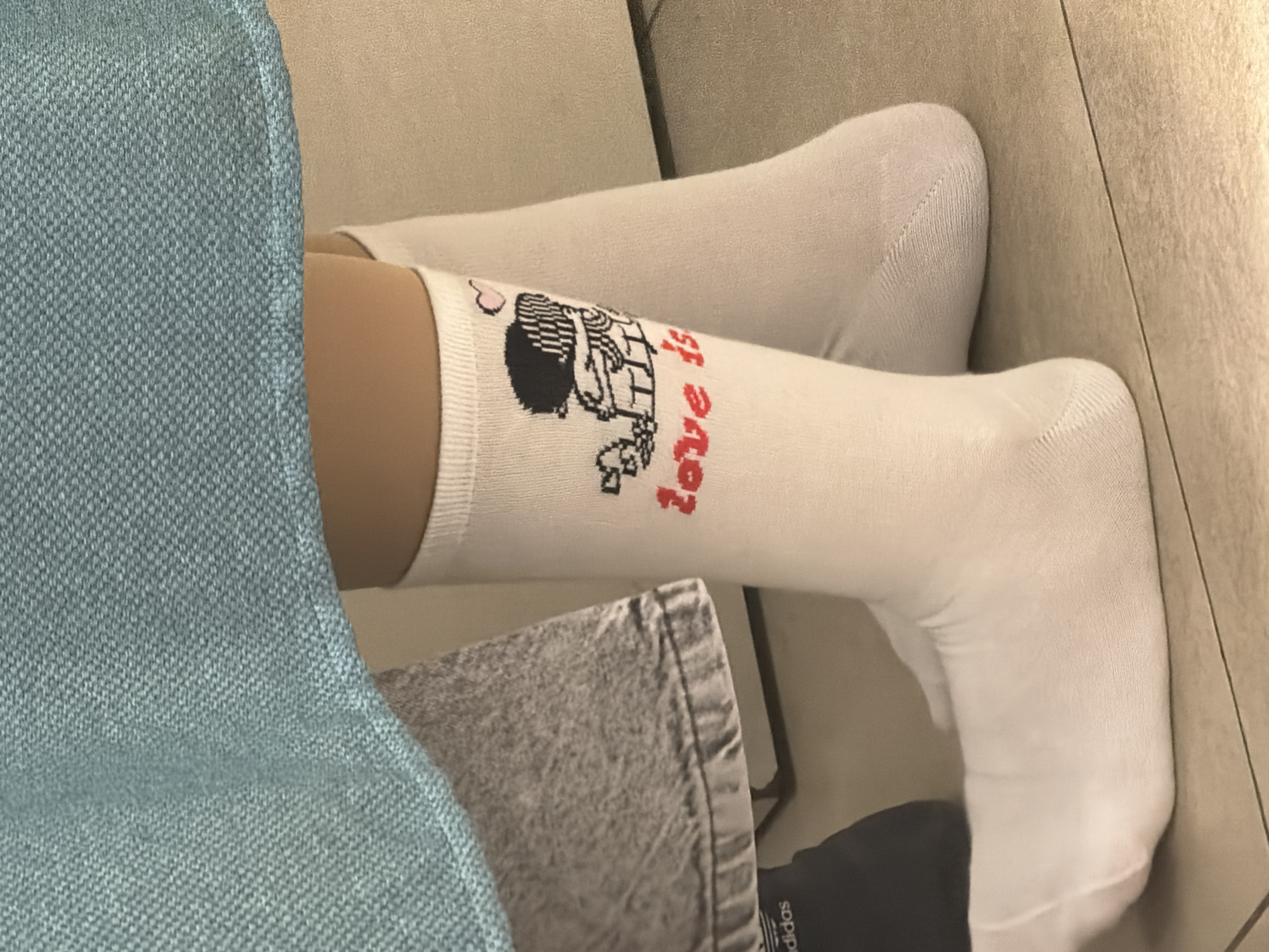 The image shows a person wearing white knee-high socks. One sock has a cartoon illustration of two figures and the words "love is" in red. The person appears to be sitting on a light-colored couch or cushion. A portion of blue fabric is visible on the left side of the image, partially obscuring the scene. A piece of gray fabric is also visible, and a black Adidas shoe can be seen near the bottom of the image. The background consists of a wall with a light-colored texture. The location in the city of Moscow, Russia cannot be determined from the image.