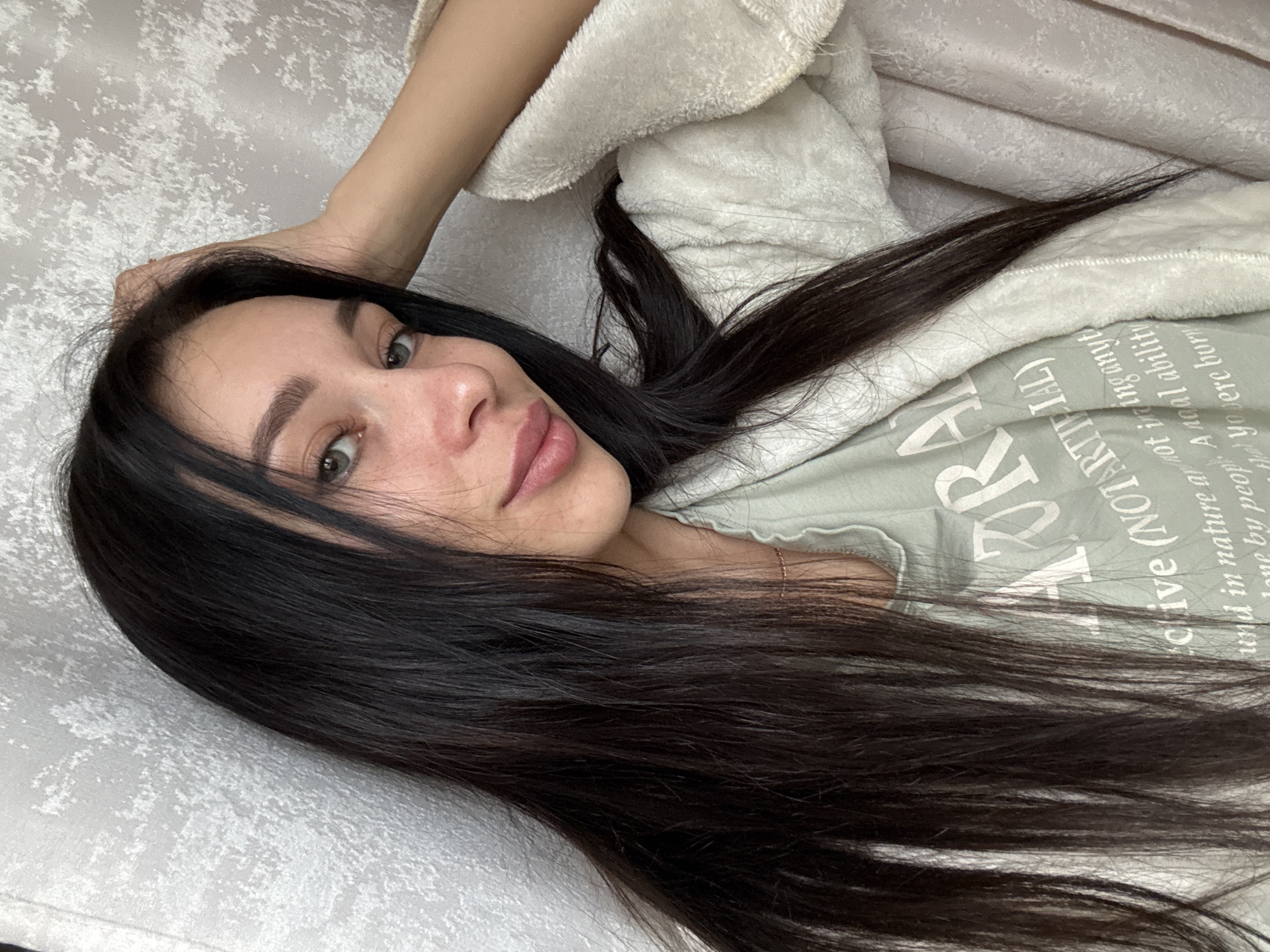 A woman with long, dark hair is lying on a patterned grey surface. She is wearing a light green shirt with white lettering and a white robe. Her face is visible, showing her eyes, nose, and lips. The lettering on her shirt contains the phrase "chive (NOT ARTIFIAL)," with the rest of the text less clear but appearing to include words like "nature" and "ability." The image appears to be taken indoors. There is no verifiable evidence in the image to suggest this was taken in Chaikovsky, Russia.