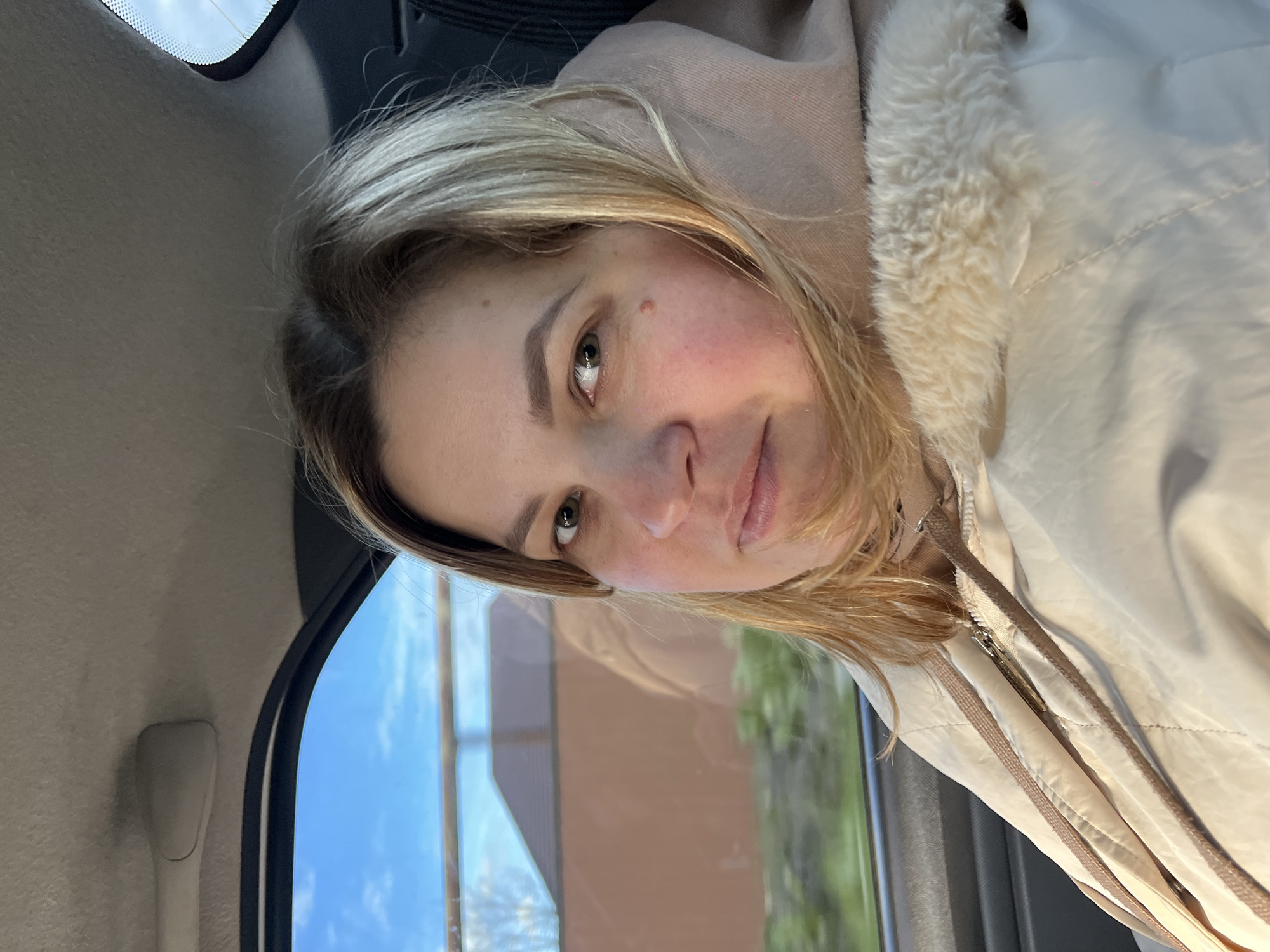A fair-skinned woman with blonde hair is sitting inside a car. She has a mole on her cheek. She is wearing a light-colored jacket with a fur collar. The car window shows a view of a building and greenery outside. The exact location in the city of Novopokrovskaya, Russia, cannot be determined from the image.