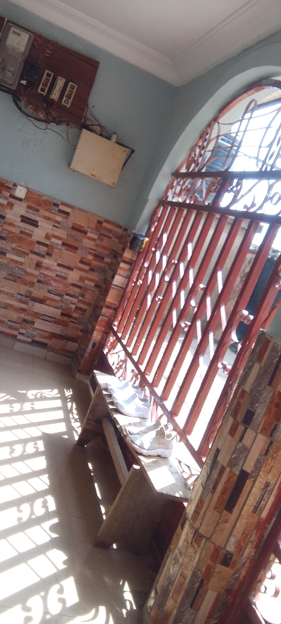 A wide shot captures an interior hallway with a light blue wall, tiled wall, and a tiled floor. To the left, an electrical meter and fuse box are mounted on a wooden board attached to the wall. A beige electrical box is mounted below the fuse box. In the center, a wooden bench is placed in front of a red metal gate. Two pairs of athletic shoes are placed on the bench, one white pair in the back and a patterned pair in the front. The gate has intricate scrollwork and is partially open, allowing sunlight to cast shadows on the floor. The ceiling features white crown molding.