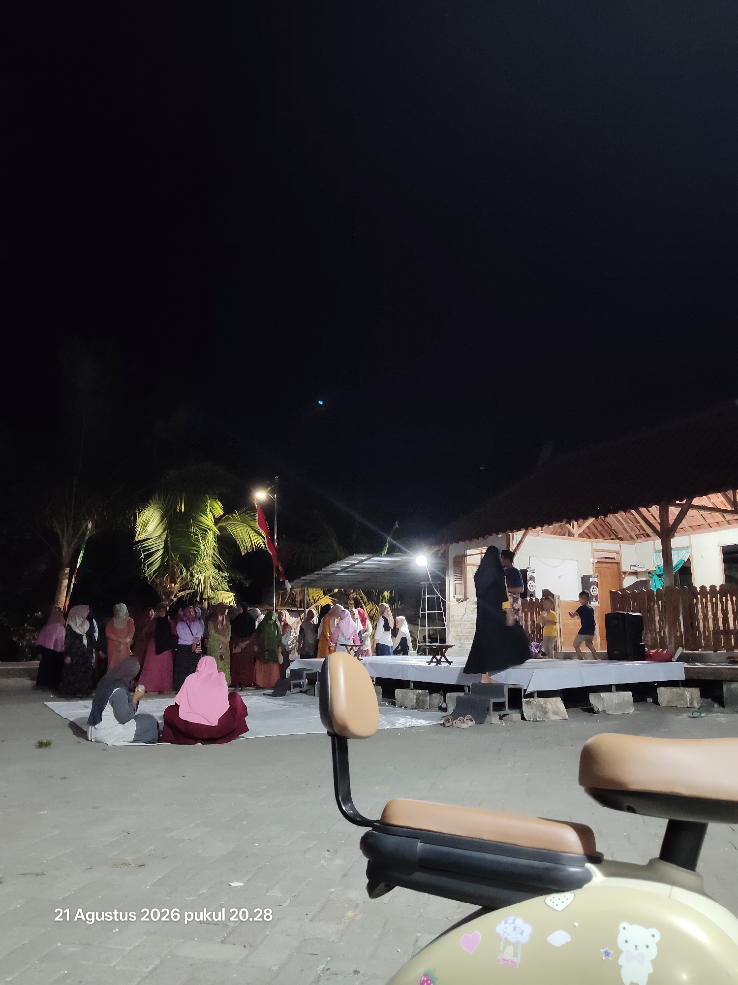 The image captures an outdoor event at night in Wedi, Indonesia. A group of people, primarily women wearing hijabs and traditional clothing, are gathered in front of a stage. On the stage, a woman in a black abaya is interacting with two children. The background features a building with a thatched roof, palm trees illuminated by lights, and a flagpole with a red and white flag. The foreground shows a portion of a beige vehicle, possibly a scooter, with stickers on its body. The ground appears to be paved with gray stones. The timestamp "21 Agustus 2026 pukul 20.28" is visible in the bottom left corner, indicating the date and time the photo was taken.