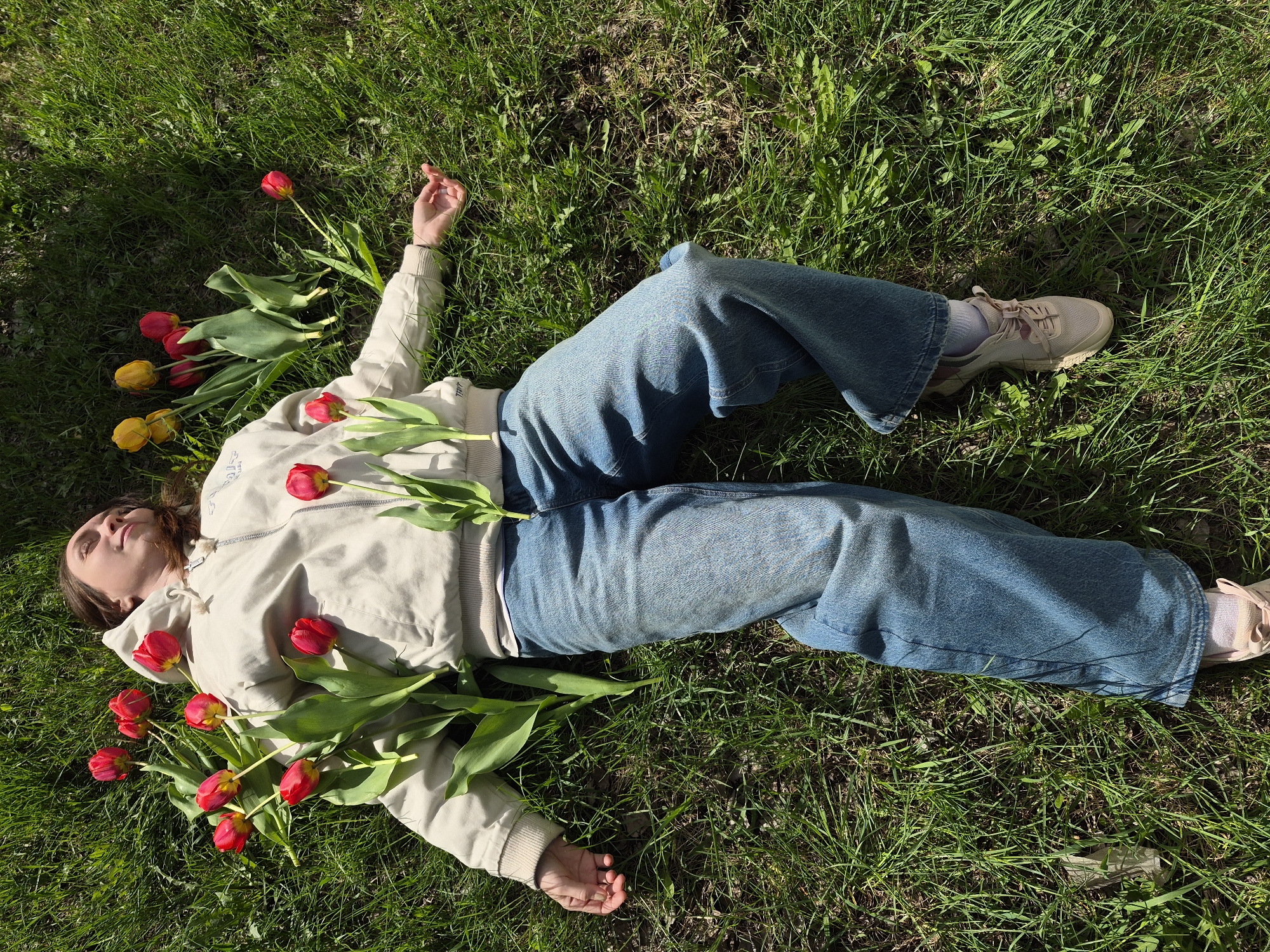 A person is lying on a grass lawn, surrounded by tulips. The person is wearing blue jeans, a beige jacket, and light-colored sneakers. They are lying on their back with their arms outstretched. Several red and yellow tulips are positioned around their head and torso. The environment appears to be a grassy area, possibly a park. There is no information in the image that would allow me to determine the location of the image within the city of Chaikovsky, Russia.