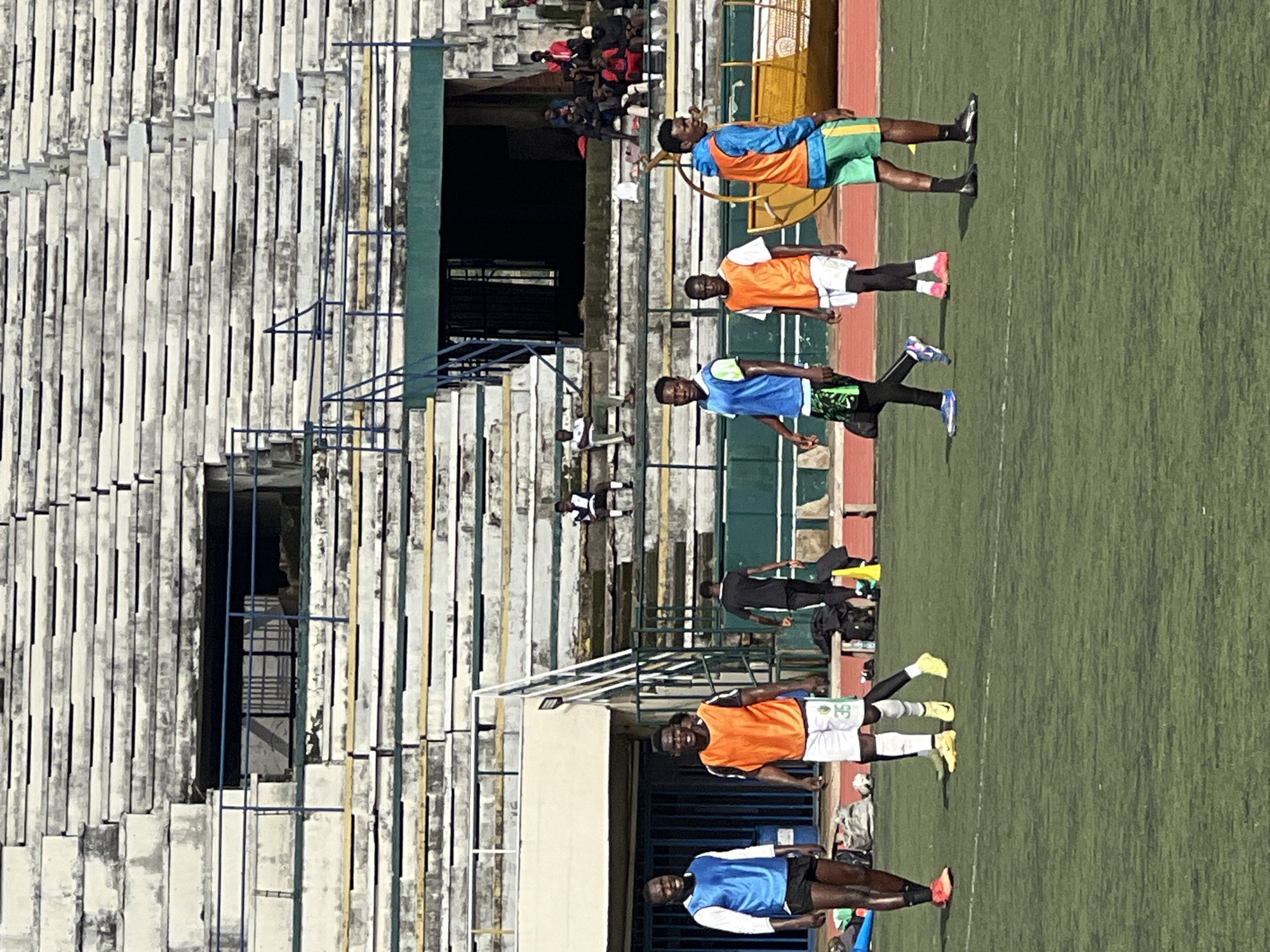 A group of young men in athletic attire are gathered on a green turf field, likely a soccer field, in front of a stadium with tiered seating. The stadium appears weathered, with some areas showing signs of disrepair and staining on the concrete seats. Several individuals are wearing training bibs over their t-shirts, suggesting a practice or training session is underway. Two individuals are prominently positioned in the foreground, one in a blue bib and the other in an orange bib. Other people are visible in the background, some seated in the stadium stands and others on the field. The overall setting is an outdoor sports facility.