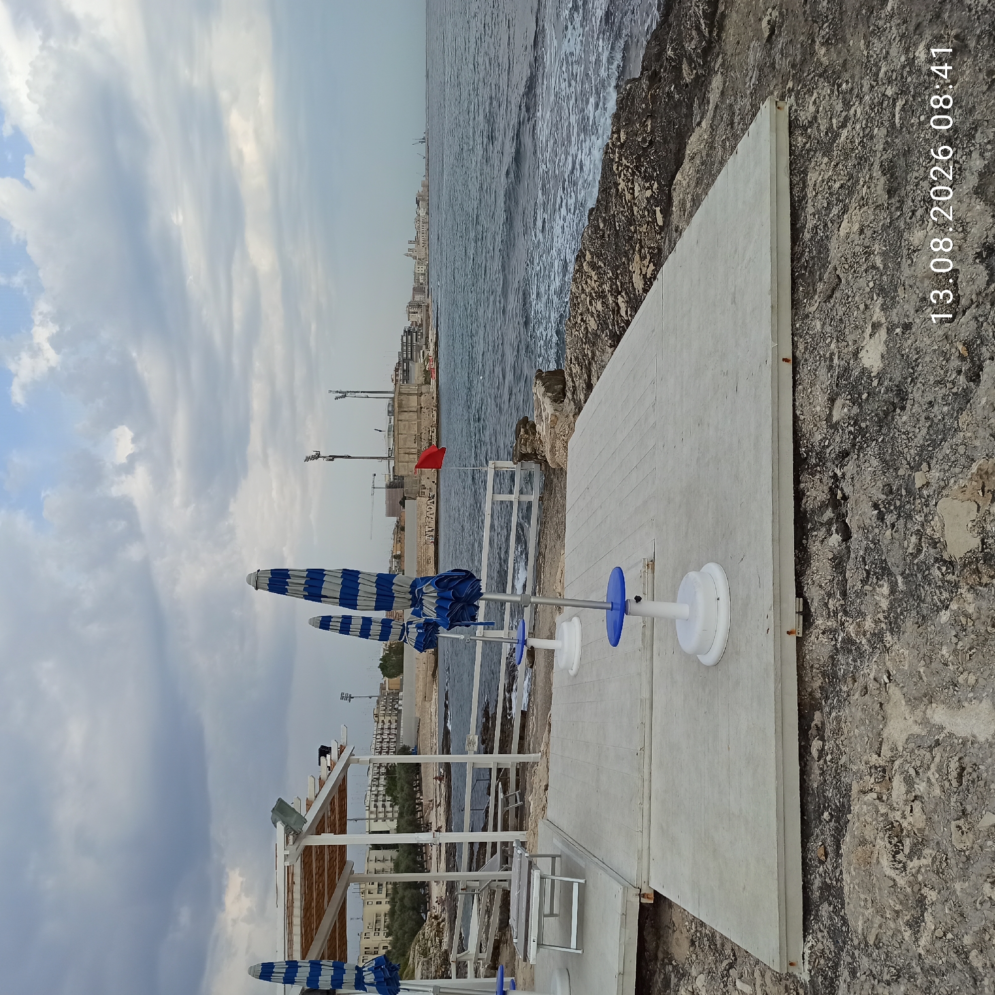 The image shows a cloudy day at a beach in Molfetta, Italy. The foreground features a raised wooden deck with several closed beach umbrellas and lounge chairs. The umbrellas are blue and white striped, and the lounge chairs are grey. A red flag is visible in the distance, near the water. The sea is choppy, and there are rocks along the shoreline. In the background, there are buildings and tall lights, possibly for a sports stadium. The timestamp on the image reads "13.08.2026 08:41". There are no people clearly visible in the immediate foreground, but a few are scattered on the beach in the middle ground. The overall impression is of an empty, overcast beach.