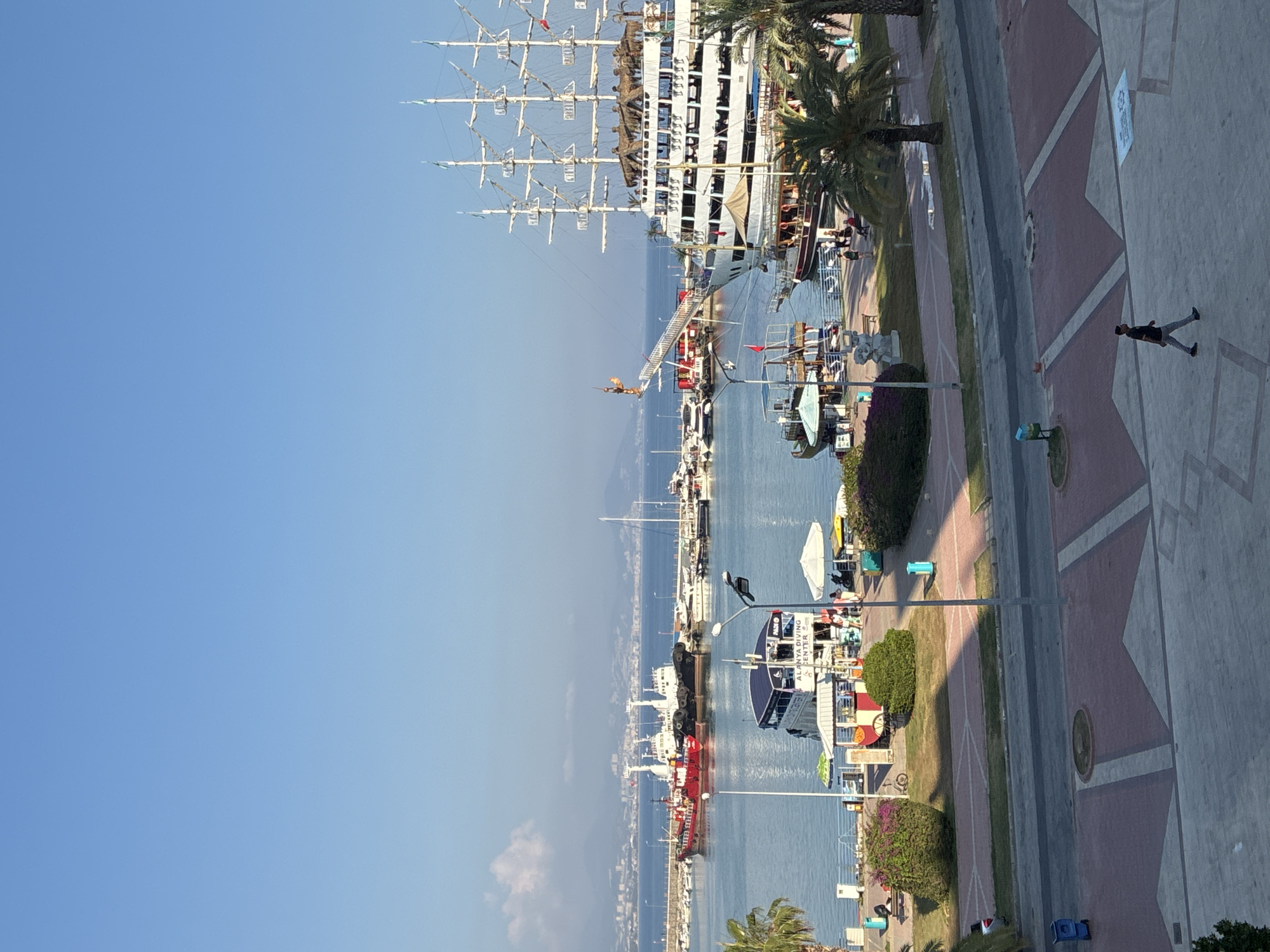 An aerial shot captures a scenic view of Alanya's harbor in Turkey. The clear blue sky stretches above a bustling marina filled with various boats, including a large, white sailing ship with multiple masts and a red cargo vessel. Palm trees line the waterfront promenade, which is paved with red and gray tiles. People are seen walking along the promenade and on the boats. The water is calm, reflecting the bright sunlight. In the foreground, a lone figure walks across the tiled pavement. The city's skyline is visible in the distance across the water, with mountains forming a backdrop.