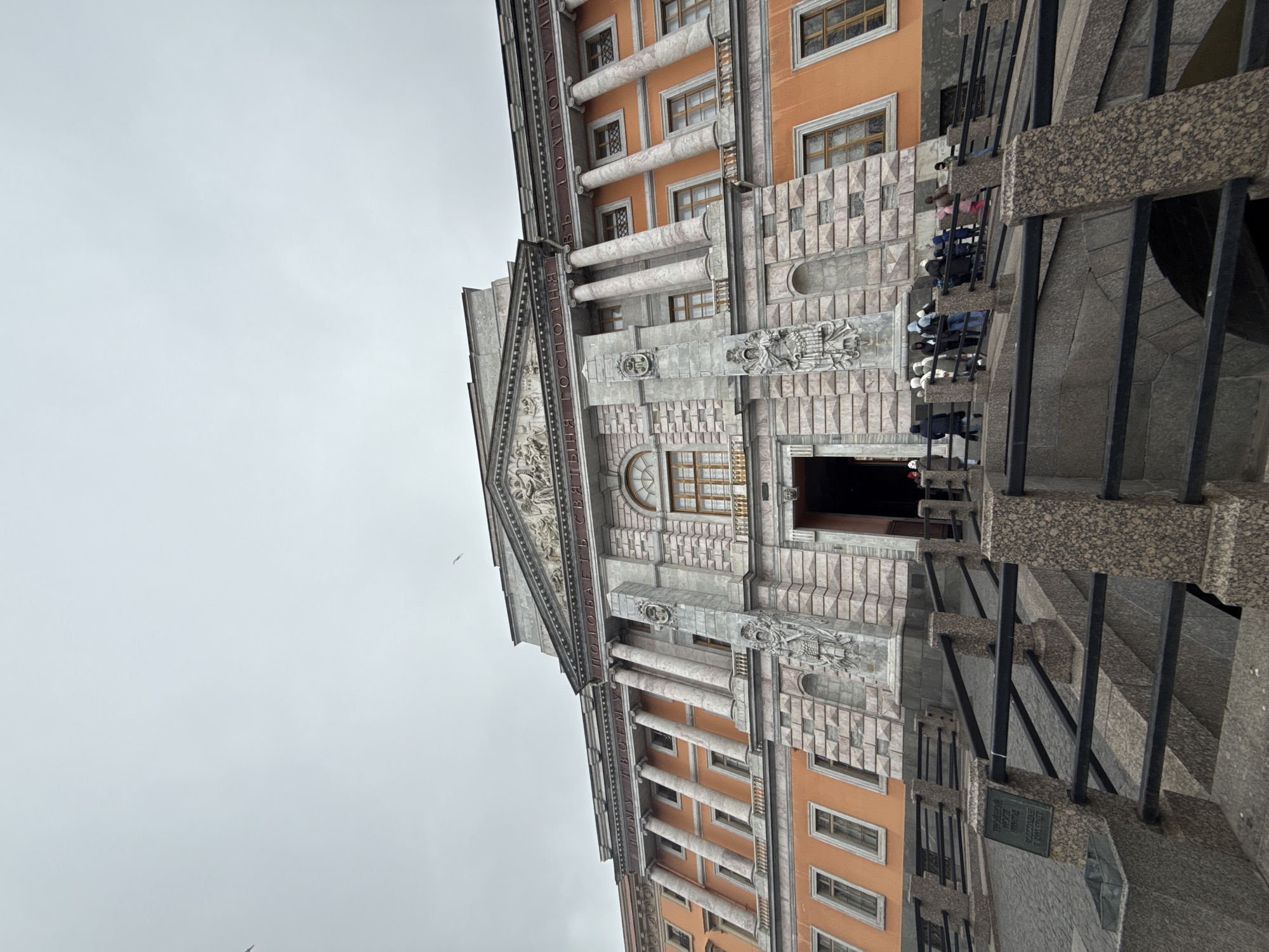 The image displays a grand, ornate building, the Mikhailovsky Castle (also known as St. Michael's Castle), located in Saint Petersburg, Russia, under an overcast sky. The photograph is oriented vertically, showing the building rotated counter-clockwise.
The primary subject is the building's elaborate facade, showcasing classical architecture. It features a prominent central section with a large pediment adorned with detailed sculptural reliefs. Below the pediment, a large arched window with a grid pattern is visible above what appears to be the main entrance. The building's exterior combines orange-colored walls with extensive grey stone elements, including numerous columns with fluted shafts and decorative capitals, and textured stonework. Multiple rectangular windows with intricate grilles are visible across the facade.
A group of people, appearing to be visitors or tourists, are present on a set of stone stairs or a viewing platform to the right of the main entrance. They are casually dressed, suggesting a daytime visit. Dark metal railings and speckled granite steps lead up towards the building.
The weather is overcast, indicated by the uniform grey sky, suggesting a cool or mild day. No specific time of day can be determined beyond daylight.
Notable visible text, prominently displayed on the building's pediment, is in Cyrillic: "СВЯТЫНЯ ГОСПОДНЯ ПОДОБАЕТЪ СВЯТЫНЕ НА ДОЛГОТУ ДНЕЙ" (Svyatynya Gospodnya podobayet svyatyni na dolgotu dney), which translates to "To Thy House Becomes Holiness; For Length of Days." Another small plaque is visible near the railings, but its text is illegible. A small bird can be seen flying in the sky.