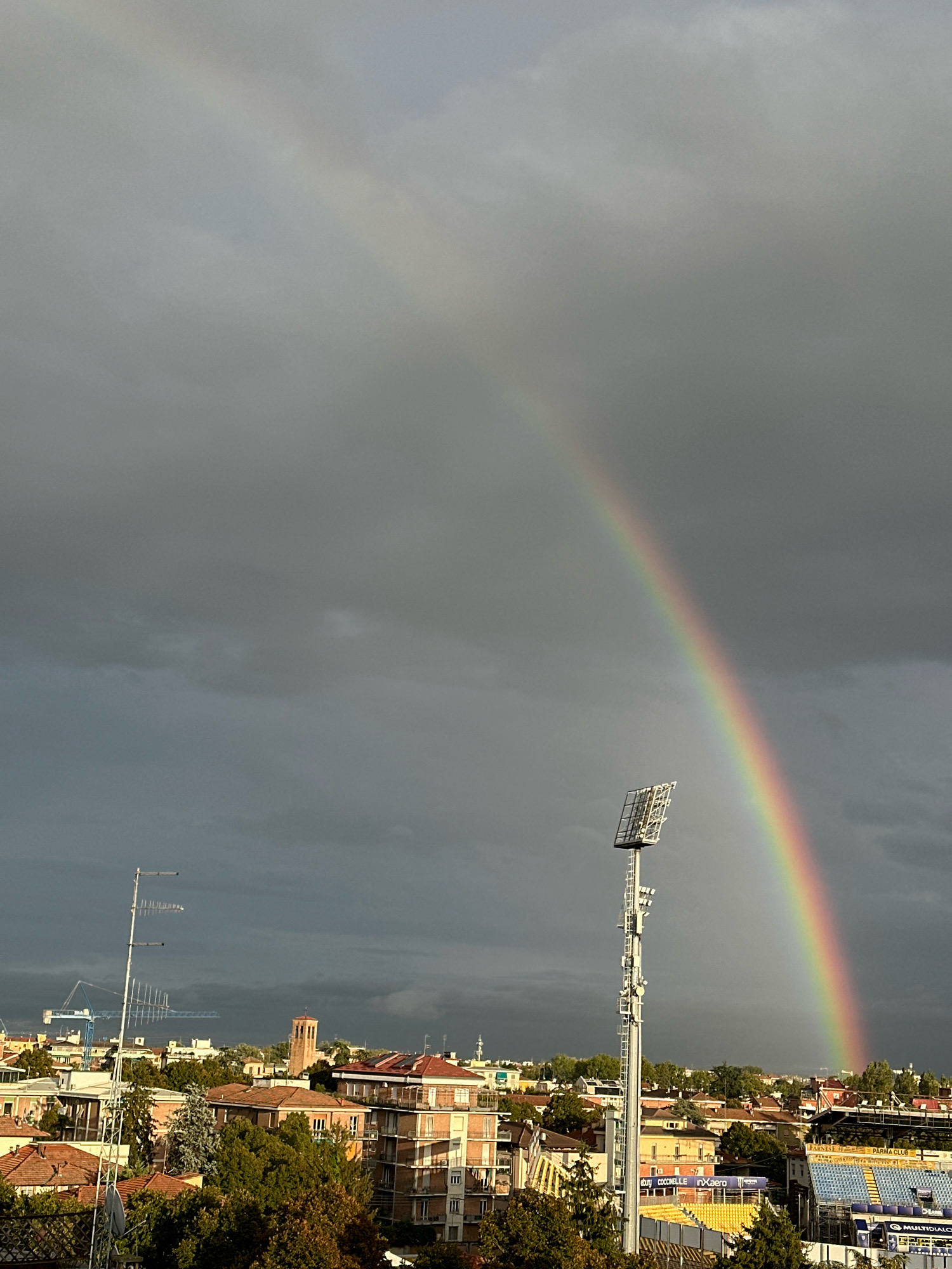 This is a vertical, eye-level, medium shot of a city skyline under a dark, cloudy sky, with a rainbow arching across the upper half of the frame. The city below is comprised of buildings with terracotta roofs, apartment buildings, a stadium with blue seating, a tall floodlight tower, a construction crane, and several antennas. The rainbow, with its distinct bands of color, appears to originate from the left side of the frame and curve towards the right. The sky is predominantly gray, with patches of lighter gray and dark clouds indicating potential rainfall. The lighting suggests it is daytime, possibly late afternoon or early morning, given the dramatic cloud cover and the visible rainbow. The scene depicts an urban environment, likely within Parma, Italy, judging by the presence of the stadium signage that reads "PARMA CLUB" and "PARMALAT", which is a well-known company with historical ties to Parma. There are no people clearly visible in the image.