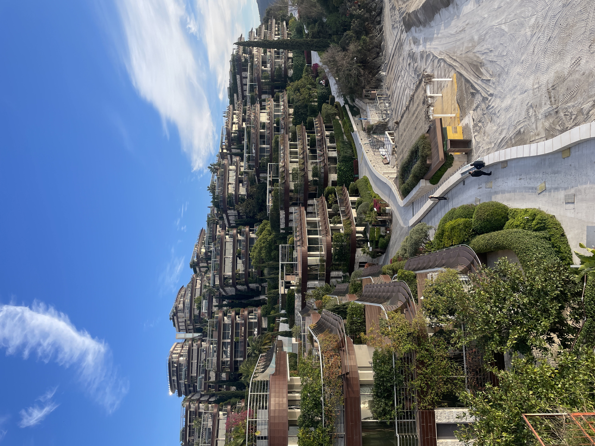 The image displays the Dukley Gardens luxury residential and resort complex located on the Zavala Peninsula in Budva, Montenegro. The scene is dominated by a hillside densely covered with multi-tiered buildings featuring modern, organic architecture with brown-toned facades, expansive balconies with white balustrades, and extensive, integrated greenery. Lush, manicured landscaping, including tall slender cypress trees, various shrubs, and flowering plants, is evident throughout the terraces and grounds.
A wide, light-colored paved pedestrian promenade, constructed from stone or concrete, curves along the base of the resort complex. This promenade adjoins a wide, sandy beach area marked by numerous vehicle tire tracks, suggesting ongoing construction or seasonal preparation. On the beach, near the promenade, an open-sided cabana with a thatched roof and white support posts is visible, alongside a smaller, enclosed wooden utility structure.
Three adult individuals are present on the promenade in the mid-ground. Two individuals are positioned close to the beach structures, one standing and the other crouching or kneeling, appearing to inspect or interact with the immediate beachfront area. A third individual is walking on the promenade, moving away from this pair. The sky above is clear blue with several scattered wispy white clouds. The natural illumination suggests soft daylight, possibly morning or late afternoon.