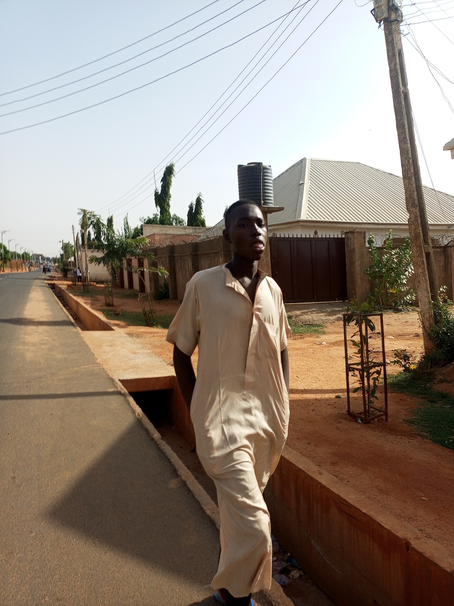 A young man stands on a street in Tempure, Nigeria, near a shallow concrete drainage ditch separating the paved road from a dirt shoulder. He wears a light-colored, traditional garment. Overhead, power lines stretch across the sky. In the background, buildings with metal roofs and a water tank are visible behind a wall.