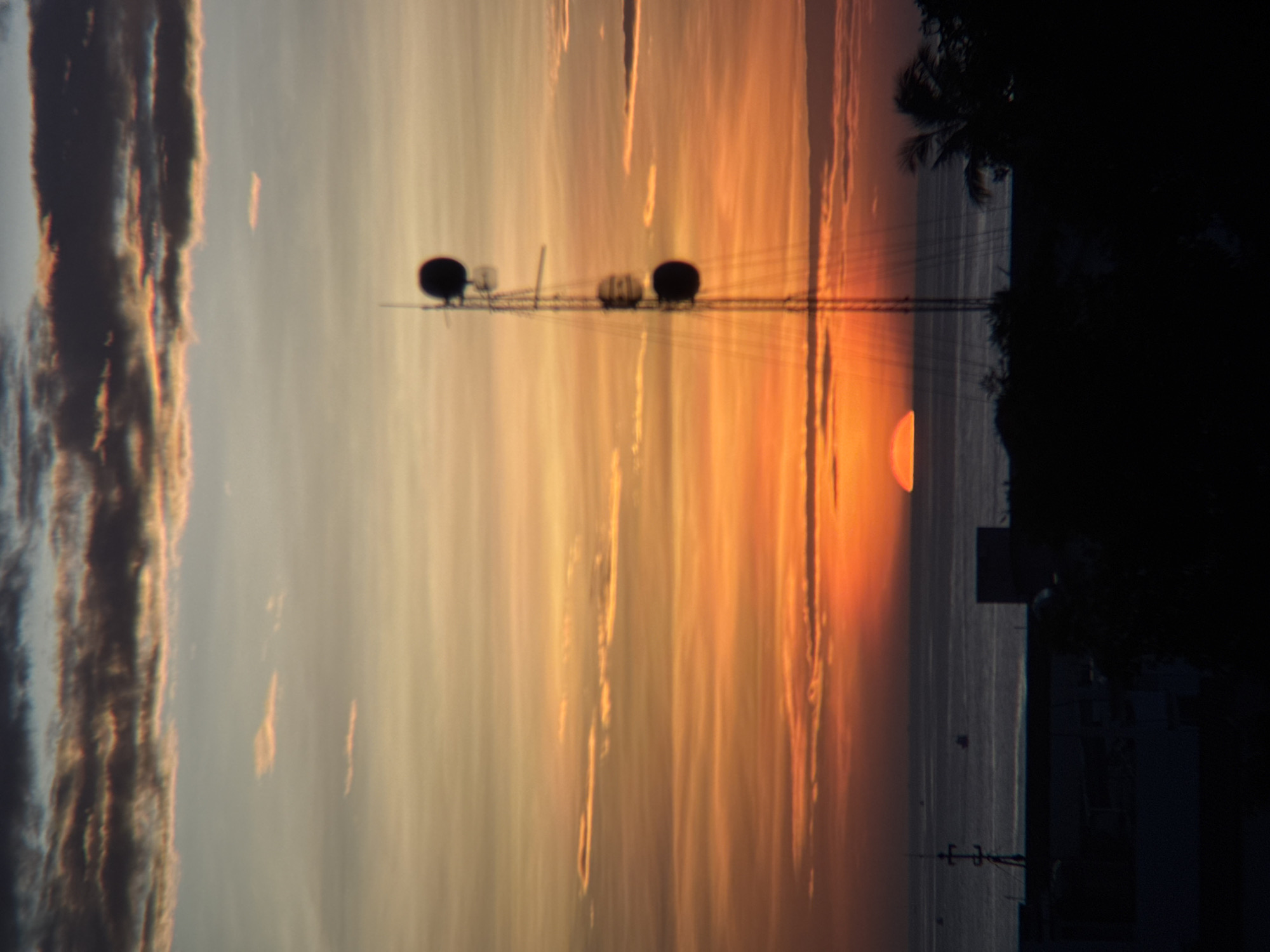 The image depicts a sunset over the ocean in Nha Trang, Vietnam. The sky is filled with vibrant hues of orange, yellow, and pink, with clouds streaking across the horizon. A silhouetted communication tower stands prominently on the right side of the frame, its structure casting a long shadow. In the foreground, the dark silhouettes of buildings and trees are visible, suggesting an urban or coastal setting. The sun, a bright orange semi-circle, is partially submerged below the ocean's surface, casting a warm glow across the water. Several small boats are scattered across the sea, adding a sense of scale to the vast expanse of water. The overall atmosphere is serene and tranquil, capturing the beauty of a tropical sunset.