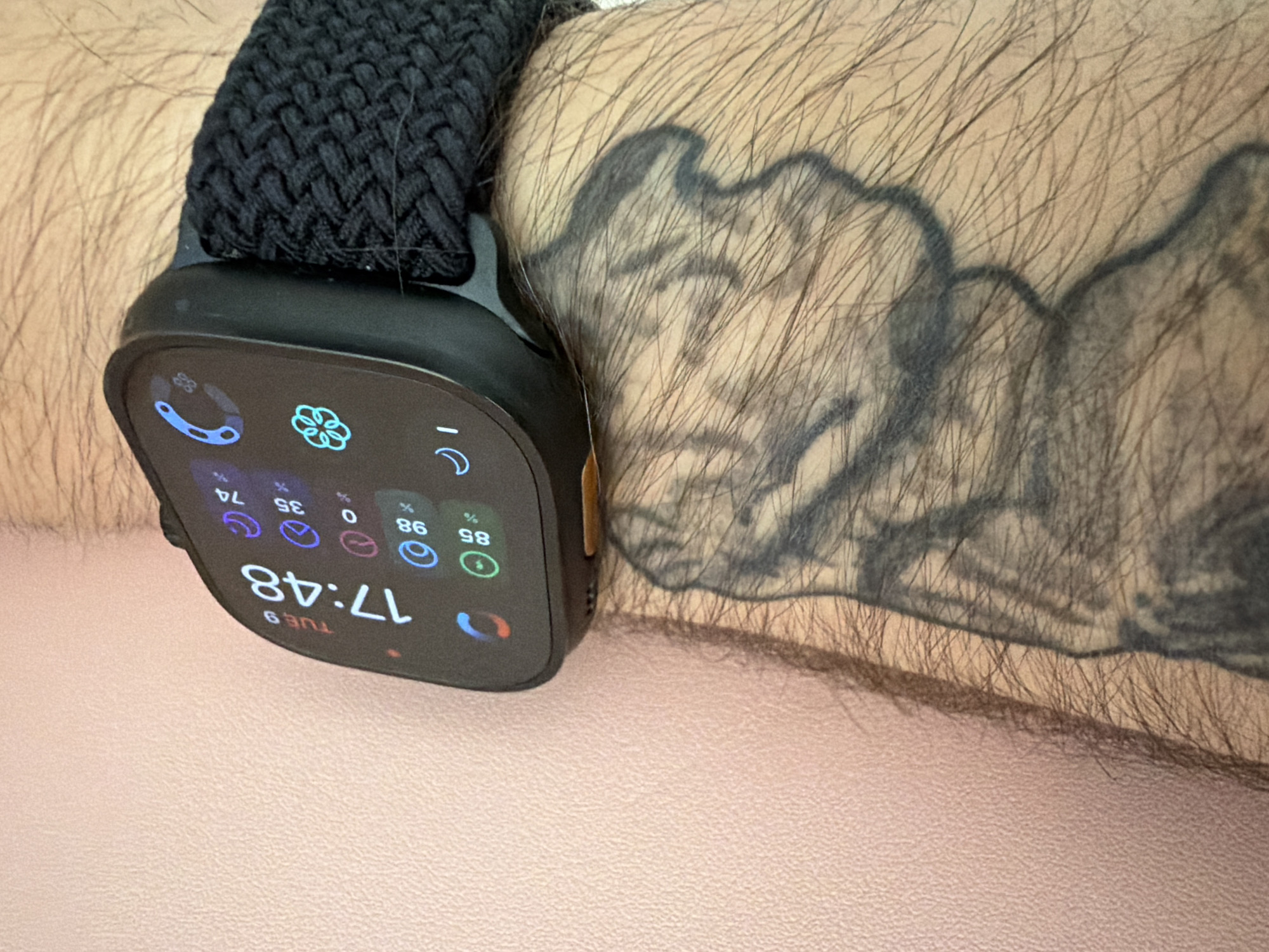 The image shows a close-up of a person's wrist wearing a black smartwatch with a braided band. The watch face displays the time as 17:48, the day as TUE 9, and various app icons and data points. The person has a tattoo on their arm, partially visible to the right of the watch. The background is a textured pink surface. There is no indication of the location being in Dobrich, Bulgaria.
