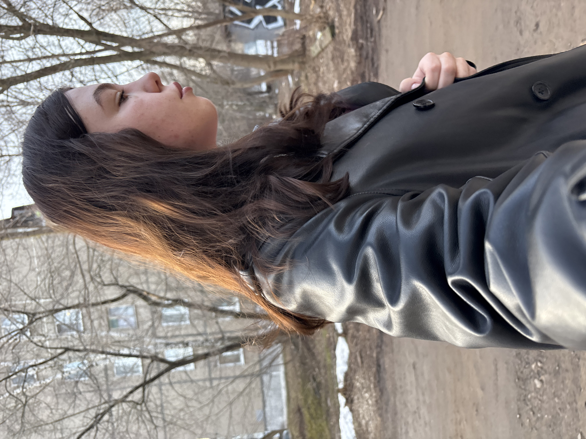 A young woman with dark hair is standing outdoors in what appears to be a city environment. She is wearing a black leather jacket and her head is tilted back, her eyes closed. In the background are bare trees, a grey building with windows, and a patch of ground. The setting is likely Perm, Russia based on the filename of the image which contains that location. The ground appears to be a dirt path or unpaved area, with some patches of what may be melting snow.