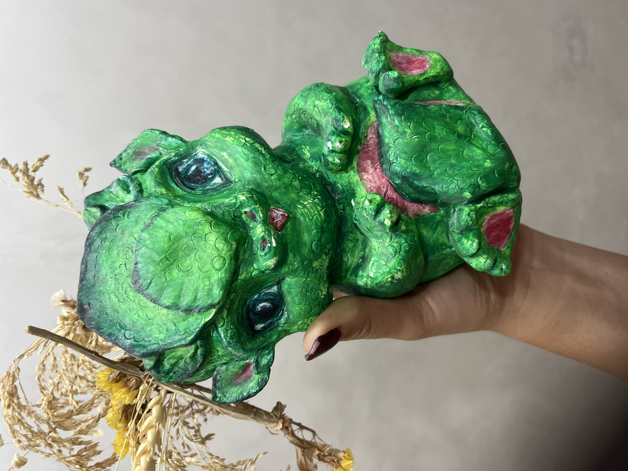 In a close-up shot, a person's hand holds a small, bright green, dragon-like figurine. The figurine has large blue eyes, pink accents on its ears and stomach, and a textured, scaled appearance. The figurine is positioned in front of a blurred background, with some dried grass and a small yellow flower visible to its left. The image appears to be taken indoors, with a plain, neutral-colored wall behind the subject. There is no discernible information about the location within Сеніцкі сельскі Савет, Belarus.