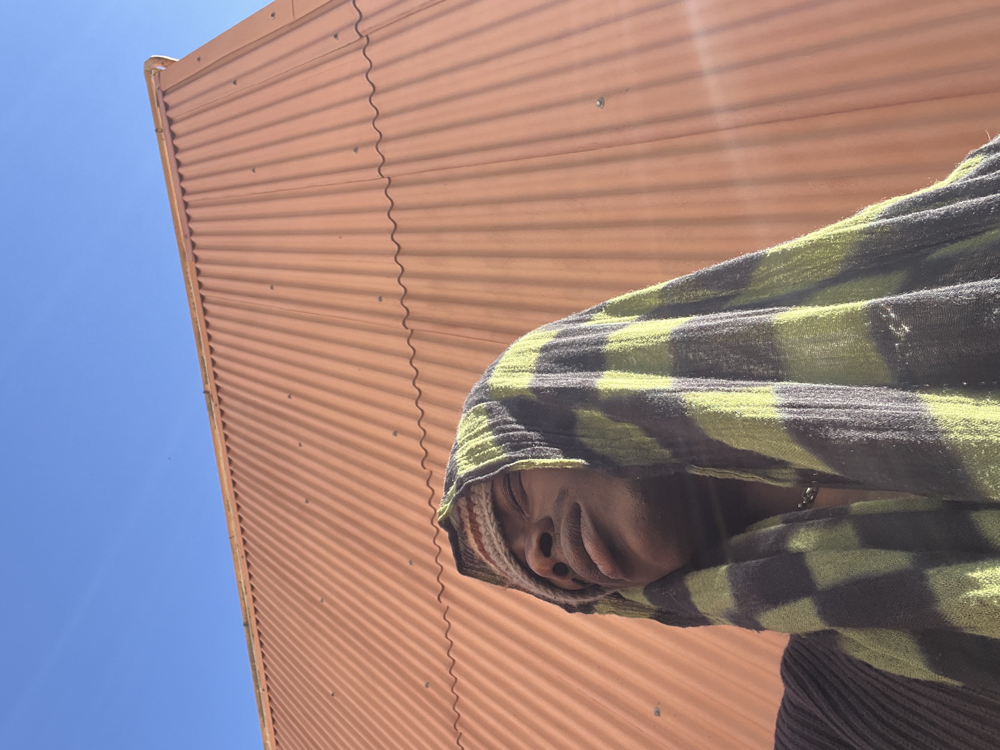 A low-angle shot shows a person partially covered with a green and black checkered hooded towel, posing against a background of a corrugated brown metal wall, possibly a building's exterior, and a bright blue sky. The towel's pattern is a mix of light green and dark grey or black checks. The person's face is mostly obscured by the towel, but part of the nose, mouth and chin are visible. The angle of the shot is tilted, making the background appear slanted. No specific evidence indicates this image was taken in Barcelona, Spain.
