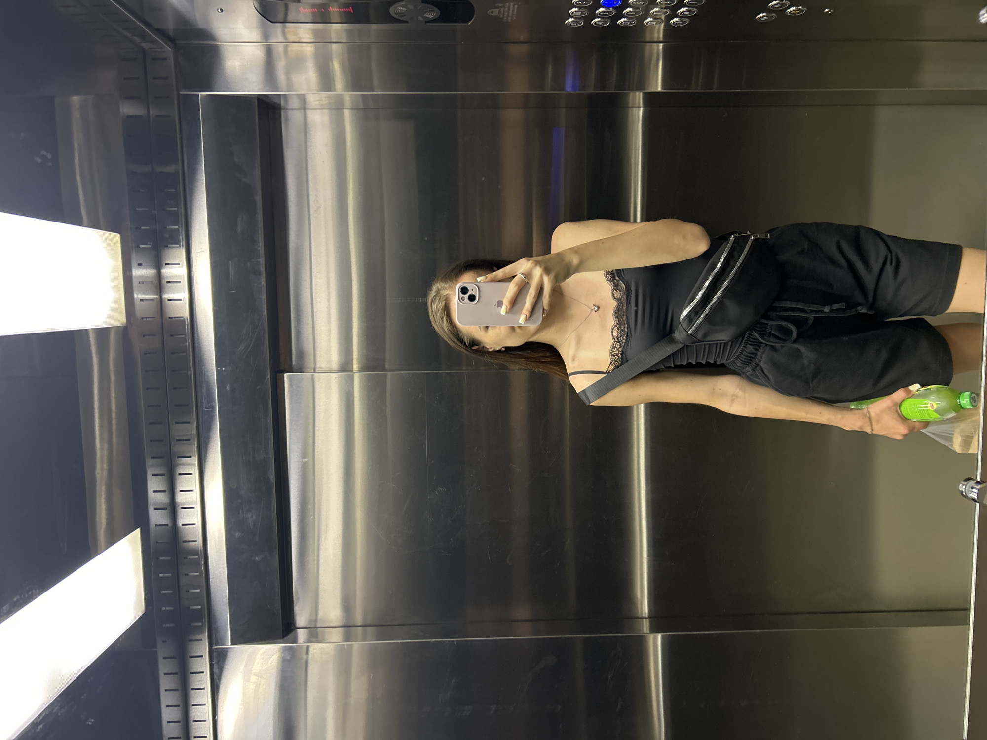 A young woman is taking a mirror selfie in an elevator. She is wearing a black tank top with lace trim, black shorts, and a black fanny pack across her chest. She is holding a green bottle in her left hand. The elevator walls are made of brushed stainless steel, and there are two fluorescent lights on the ceiling. An elevator control panel with several buttons is visible on the right side of the elevator.