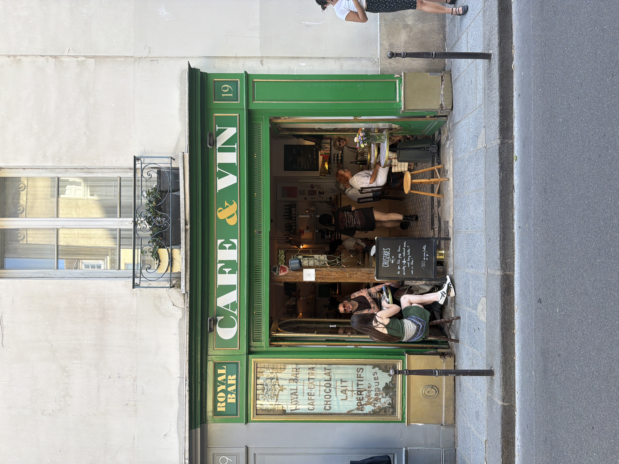 The image shows the exterior of a café in Paris, France. The cafe, named "Café & Vin," features a green and gold facade with signage including "Royal Bar," and "Café Extra." The number "19" is prominently displayed above the entrance. There is a window to the left of the entrance with a decorative wrought iron planter below.
Several people are visible: two patrons are sitting outside at the entrance with a menu board nearby. Inside the café, two additional patrons are seated at a table. A person, possibly a pedestrian, is partially visible at the top right of the frame, standing on the sidewalk. The street and sidewalk are visible in the foreground.