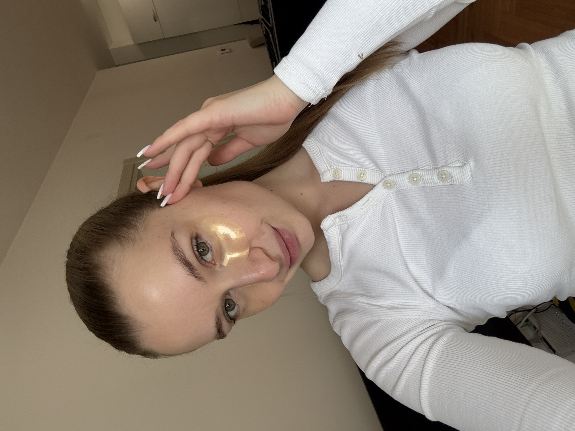 A woman wearing a white long-sleeved, button-up shirt is posing for a selfie indoors. She has light skin, green eyes, and light brown hair pulled back. She is wearing a gold eye mask under one eye. Her left hand is near her face, with long white nails. The background is a beige wall and a glimpse of a dark object. The location of the scene is not identifiable in the city of Budva, Montenegro.