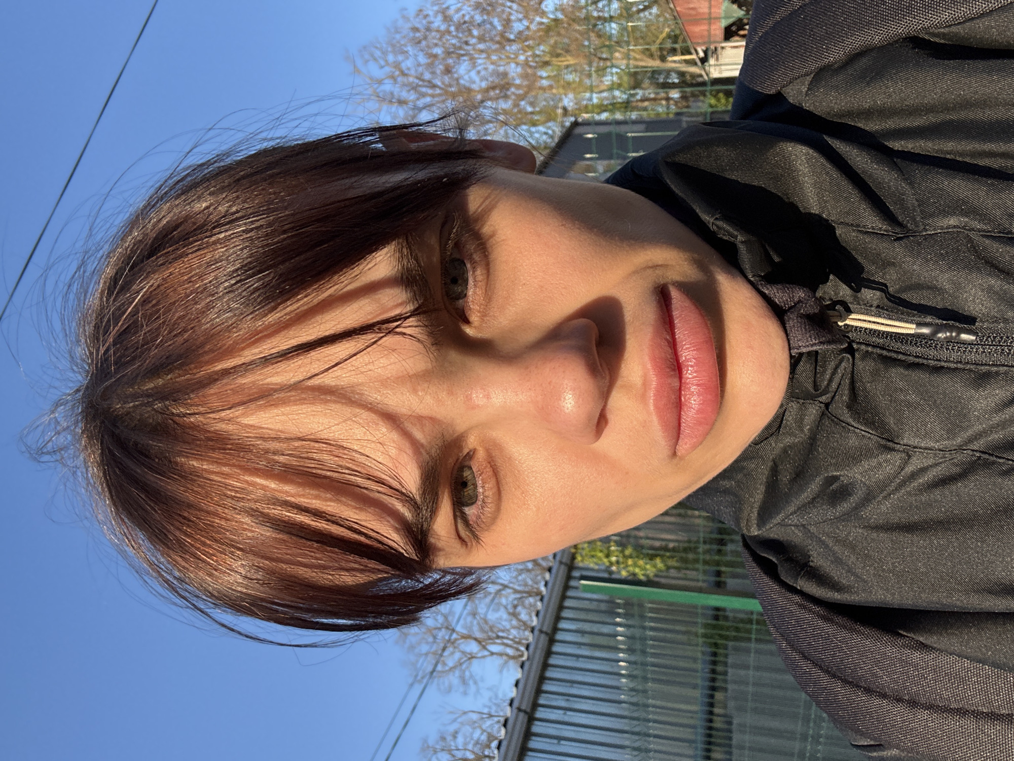 A close-up, eye-level portrait features a person, likely female, captured in a selfie outdoors in Rostov-on-Don, Russia. The person's head is tilted slightly to their right, and they look directly towards the camera with a neutral expression. They have dark brown hair, with bangs partially obscuring the forehead, and striking green or hazel eyes. Their lips are full and have a natural pink tone. Strong, direct sunlight illuminates the face from the upper right, creating distinct shadows, particularly under the nose and bangs, and highlighting the skin and hair.
The individual is wearing a dark, possibly black, casual jacket with visible fabric texture and a light-colored zipper pull tab on the right side. The background shows a clear, bright blue sky, with bare tree branches appearing on both sides, indicative of late autumn, winter, or early spring. A thin dark line, possibly a power line, crosses the sky in the upper left. In the mid-ground, a green-tinted metal fence or structure is visible. The bright, direct light signifies a sunny day. No visible text is present in the image.