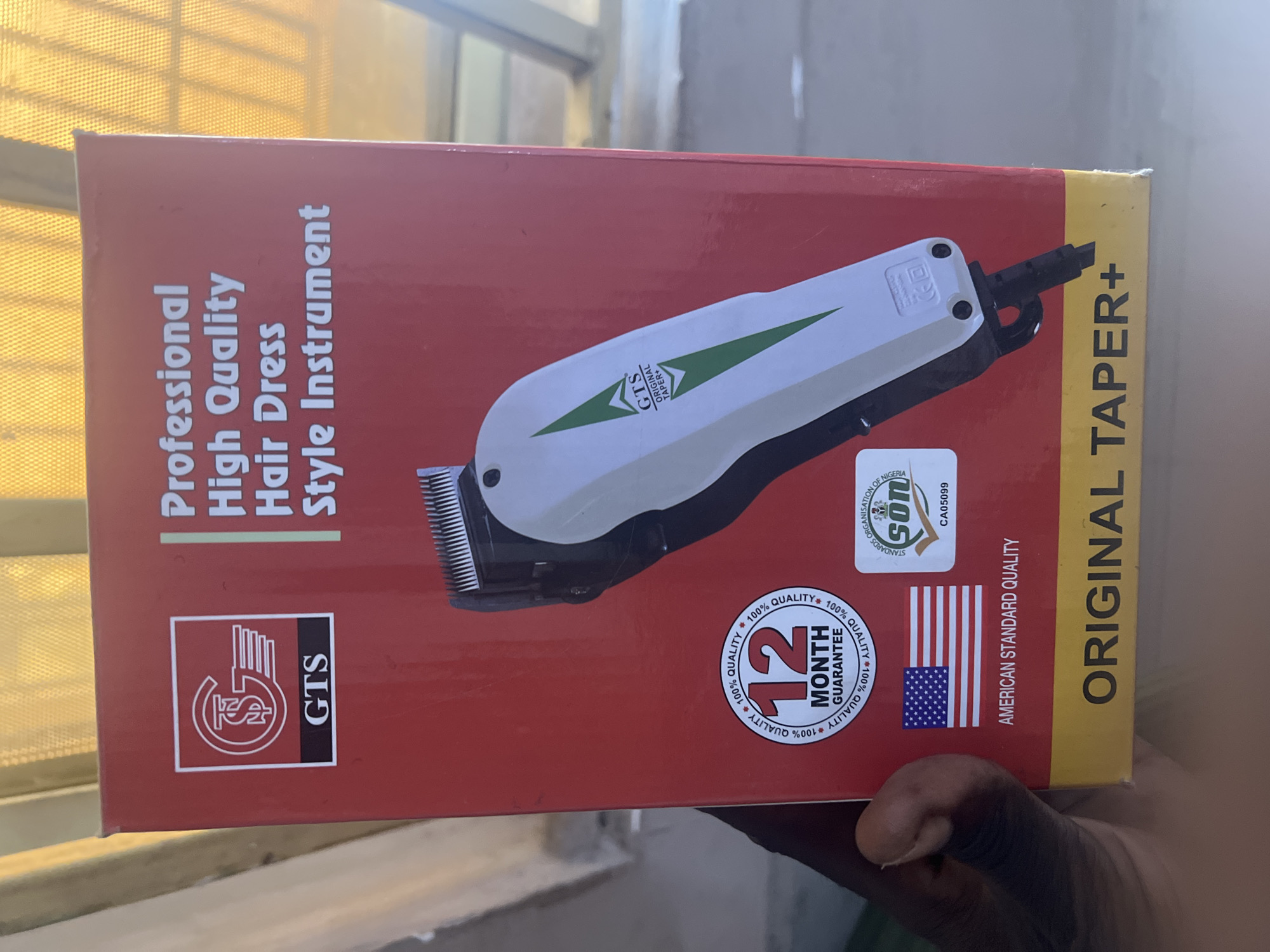 A red box for a "GTS Professional High Quality Hair Dress Style Instrument" is shown, held by a person's hand in the lower left corner. The box features the GTS logo, a depiction of the product, a "12 MONTH GUARANTEE" seal, an American flag sticker, and a SON (Standards Organisation of Nigeria) logo. The background appears to be an indoor setting with a yellow, lattice-like curtain or blinds.