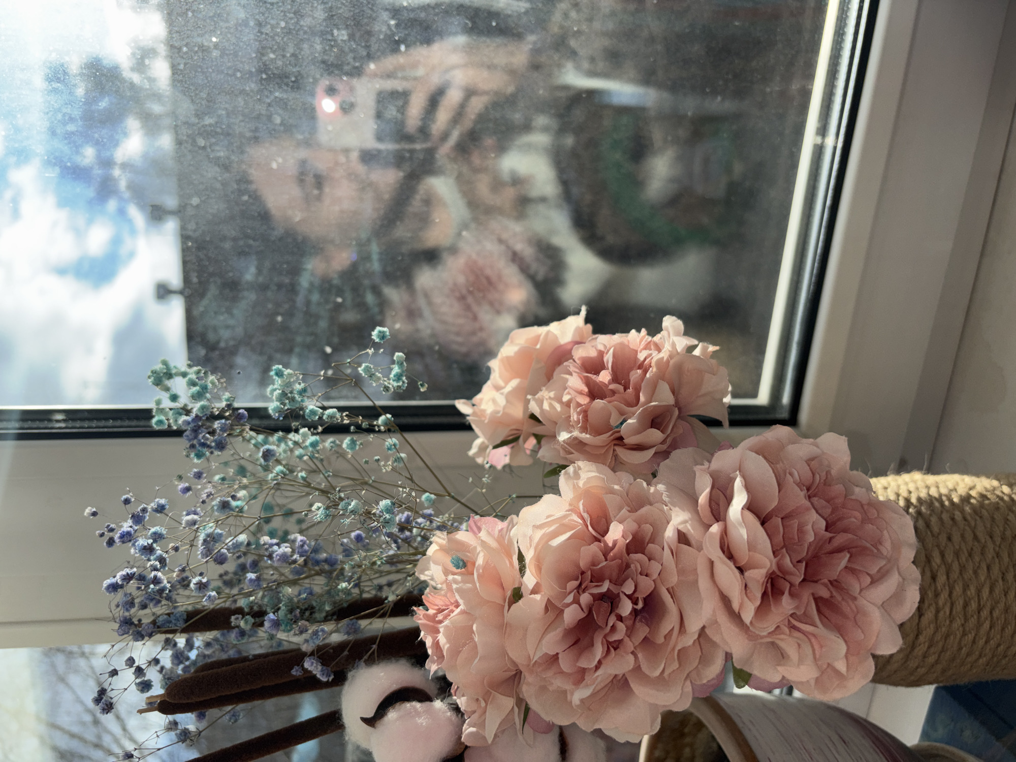 A photograph taken inside a building in Perm, Russia shows a window with flowers and other decorative objects on its sill. The window has a reflection of a person holding a smartphone. The room appears to be well-lit with natural light coming through the window.
In the foreground, there's a bouquet of pale pink peony-like flowers along with sprigs of small, light blue flowers. Also visible are decorative cattails and cotton ball decorations with black accents.
The window frame is white and modern, and the sill is also white. Beyond the window, the outside is blurred, suggesting some distance or a shallow depth of field. The reflection in the window shows a person holding a smartphone, presumably taking the picture. The reflection also reveals an area of sky with some clouds.