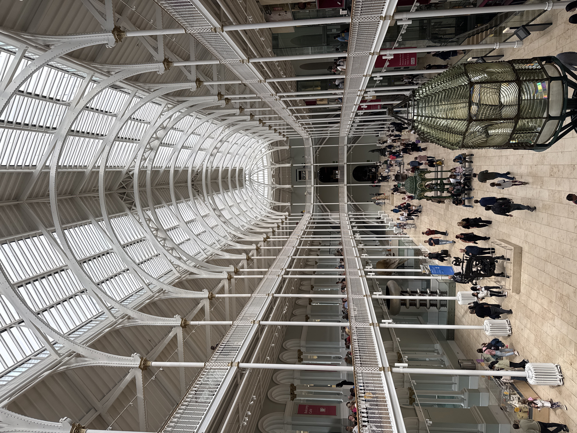 The image captures the grand interior of the National Museum of Scotland, located in the City of Edinburgh, United Kingdom. The museum features a vast, multi-level atrium with a stunning glass and iron arched roof that floods the space with natural light. Intricate white metalwork forms a series of balconies and walkways that overlook the main exhibition floor.
On the ground floor, numerous visitors are dispersed throughout the space, some walking, others observing exhibits, and a few seated on benches. Various displays and exhibits are visible, including what appears to be a large Fresnel lens in the foreground. People are seen interacting with displays, conversing, and moving between different areas of the museum. The upper levels also show visitors on the balconies, observing the scene below. The architecture is a prominent feature, with rows of columns supporting the balconies and the expansive, domed ceiling creating a sense of grandeur and openness. The overall atmosphere appears to be one of public engagement with cultural exhibits within a historically significant and architecturally impressive setting.