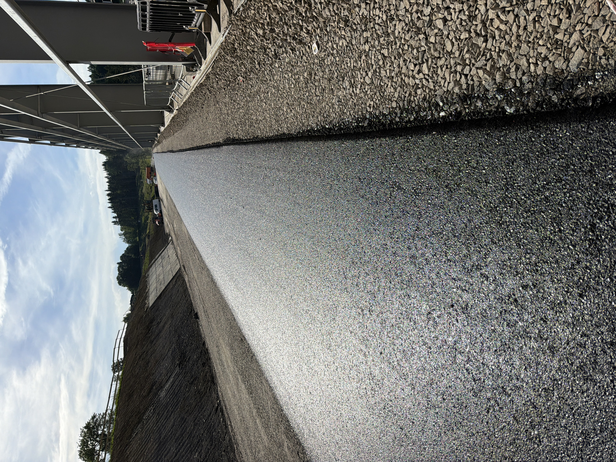 The image depicts a construction site with a freshly paved asphalt surface. The asphalt is dark gray and glistens in the sunlight, with a granular texture. To the right of the asphalt, there is a strip of coarse gravel. Beyond the gravel, a section of a large, modern-looking building with a metal frame structure is visible. Fencing and some construction equipment, including a red safety barrier and a yellow sign, are also present. In the background, a landscaped slope covered in dark soil and some vegetation leads up to a wooded area with trees. The sky is partly cloudy with blue sky visible. Further in the distance, vehicles and more construction equipment are scattered across the site. The overall scene suggests ongoing infrastructure development or renovation. The location is identified as Janowice, Poland.
