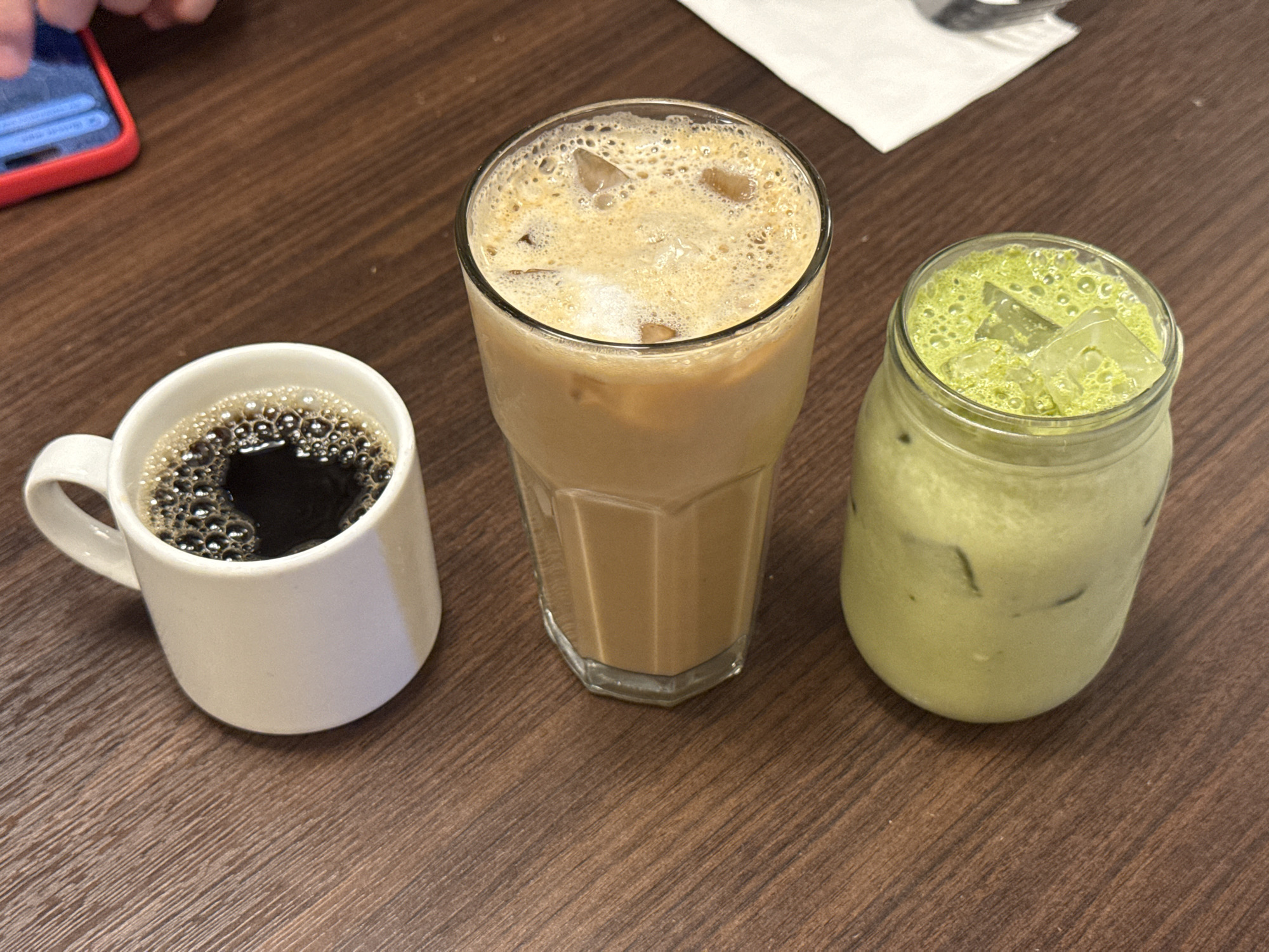 The image shows three beverages sitting on a wooden table in National City, California. On the left is a white coffee mug filled with black coffee. In the middle is a tall glass of iced coffee with a foamy top. On the right is a mason jar filled with a light green drink and ice, likely matcha. A mobile phone with a red case is visible at the top-left corner. A white napkin sits behind the iced coffee. The table is brown wood-grained.