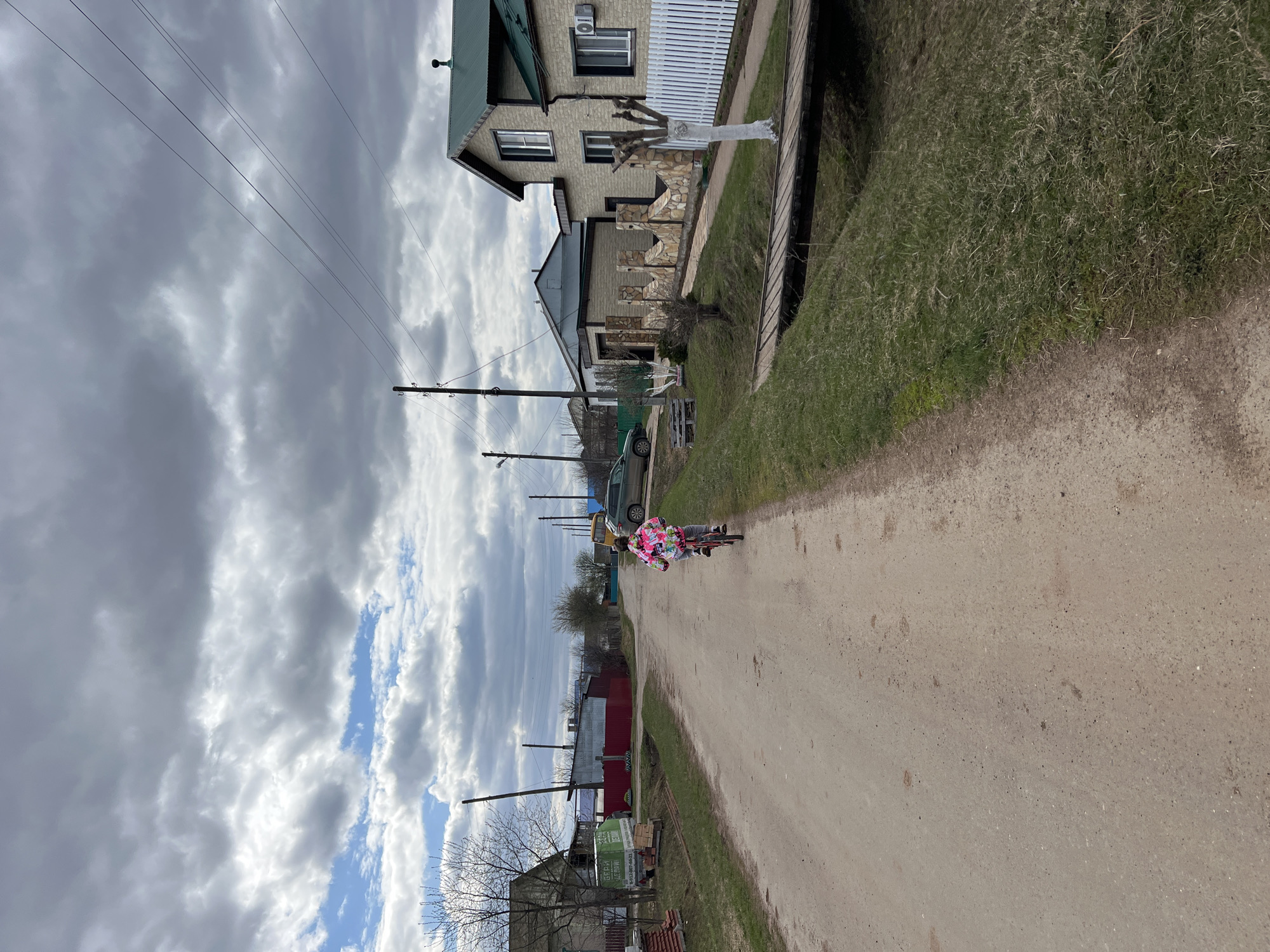 The image depicts a rural street scene in Перевозное, Russia. An individual is riding a bicycle on a dirt road, away from the camera. Houses and utility poles line the street. The sky is overcast with patches of blue. Vegetation is present along the roadside.