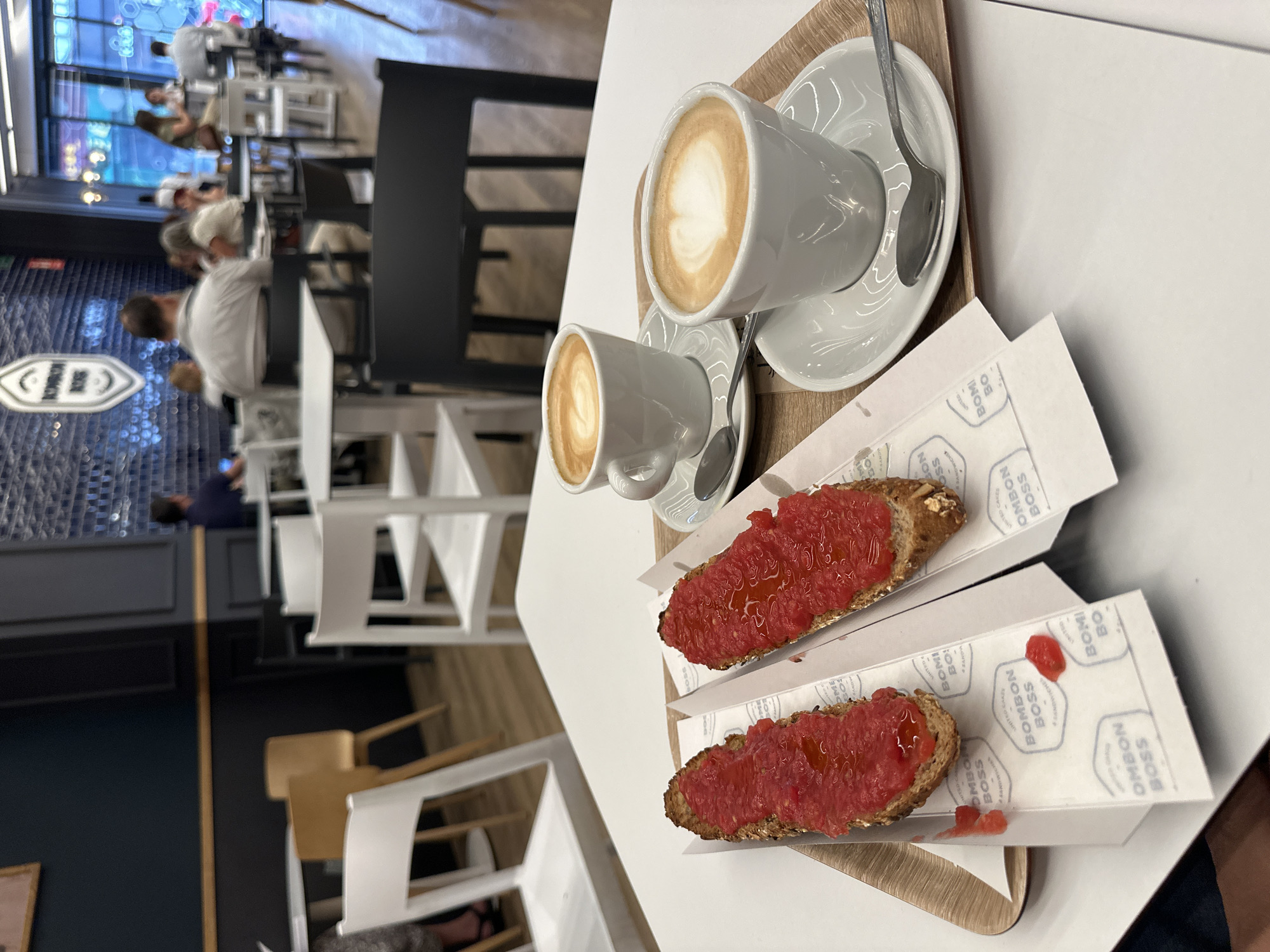 This vertically oriented photograph captures a breakfast or coffee break scene in a modern cafe. In the foreground, on a white table, a wooden tray holds two white ceramic coffee cups with saucers and spoons, each containing a latte with visible latte art. Beside the coffee, two pieces of toast, generously topped with mashed red tomato and drizzled with oil, are presented on disposable paper wrappers. These wrappers feature branding such as "BOMBON BOSS," "UNITED CAKES," "BOMI BO," and "SANDWICH."
The background shows a bustling cafe environment with several blurred figures of people sitting at tables, suggesting patrons enjoying the space. The decor includes white chairs and tables, a dark wall or counter area, and a light-colored wooden floor. A decorative, possibly metallic, sign is visible on the far left wall in the background. The lighting is bright, indicating an indoor setting, likely during daytime hours. The presence of coffee and tomato toast (pan con tomate) suggests a morning meal, possibly in a Spanish or Portuguese-speaking region, given the "Bombon Boss" brand which is known as a cafe chain. The overall atmosphere appears relaxed and casual.