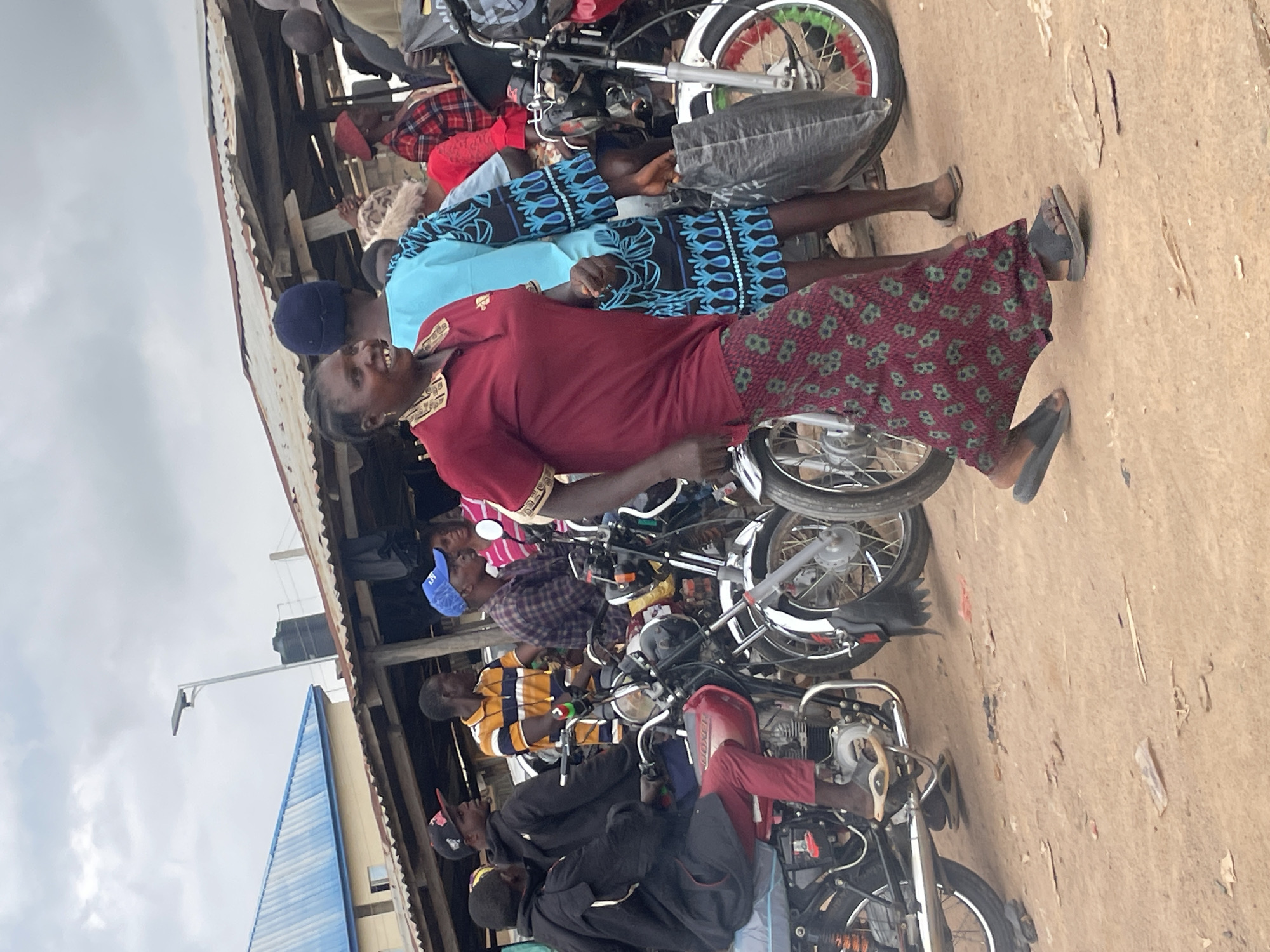 The image captures a scene in Wang, Nigeria, where a group of people are gathered around several motorcycles. The orientation of the image is rotated, as the people and objects appear to be standing vertically. In the foreground, a woman in a dark red shirt and a patterned skirt smiles, her face turned towards the viewer. Behind her, other individuals are visible, some sitting on or standing near motorcycles. The motorcycles are parked closely together, suggesting a market or a gathering point.
The setting appears to be outdoors, under a makeshift roof structure, possibly a stall or a covered area in a market. The ground is sandy or dusty. The sky in the background is overcast, indicating a cloudy day. Visible structures include a corrugated metal roof and wooden supports for the shelter. Further in the background, a tall water tank and a modern building with a blue roof are discernible, providing a contrast between traditional and contemporary elements in the environment.
The people are dressed in casual attire, with various colors and patterns evident in their clothing. Some are wearing head coverings like caps and headscarves. The overall mood seems relatively relaxed, with the smiling woman in the foreground adding a touch of warmth to the scene. The activity taking place is not explicitly defined but appears to involve people congregating or perhaps preparing for travel, given the presence of the motorcycles. No visible text is identifiable in the image.
