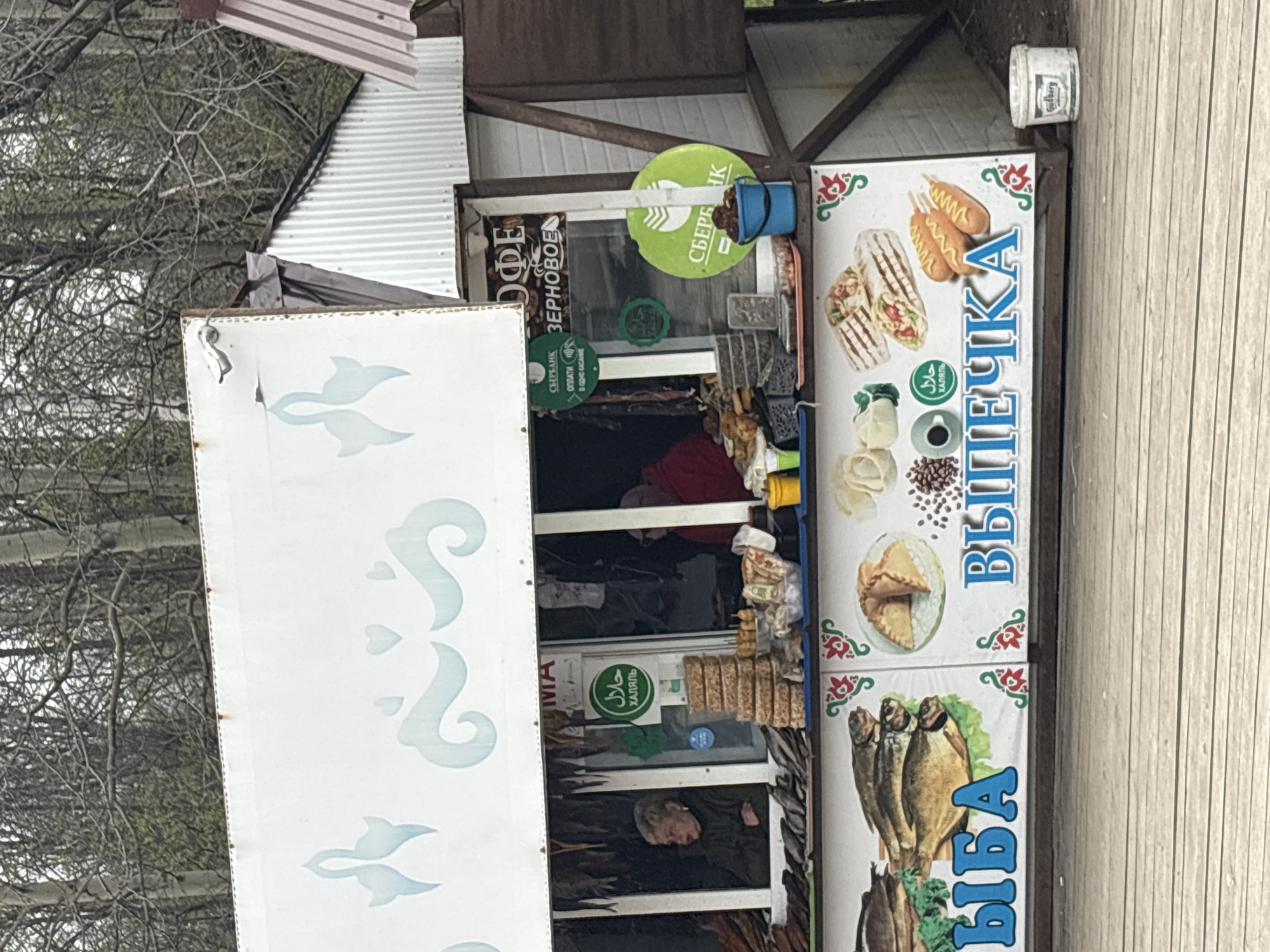 An outdoor food stall, likely a kiosk, is depicted in Nizhnie Yaki, Russia, under an overcast sky with bare tree branches visible in the background, suggesting a cooler season. The kiosk is constructed from wood and corrugated metal, with a white awning featuring light blue decorative patterns.
Inside the kiosk, a person, possibly an attendant, is partially visible, working. The stall displays various signs advertising its offerings. One prominent sign features images of shawarma/doner wraps and corn dogs, with the Cyrillic text "ВЫПЕЧКА" (Baked goods/Pastries) and a "ХАЛЯЛЬ" (Halal) logo in both Arabic and Cyrillic. Below this, another section of the sign shows images of various fish and the word "РЫБА" (Fish).
Other signs on the kiosk include "КОФЕ ЗЕРНОВОЕ" (Bean Coffee) above a window. Several green circular signs with the logo of "СБЕРБАНК" (Sberbank), a Russian bank, are displayed. One such sign reads "ОПЛАТИ В ОДНО КАСАНИЕ" (Pay in one touch), indicating contactless payment options. Another "ХАЛЯЛЬ" sign is visible near the top left inside the kiosk. A small white cup labeled "Gudberg" is placed on a wooden beam. A blue bucket hangs from a beam, and various packaged items and snacks are visible inside the stall. The overall impression is of a small, functional food and drink vendor.